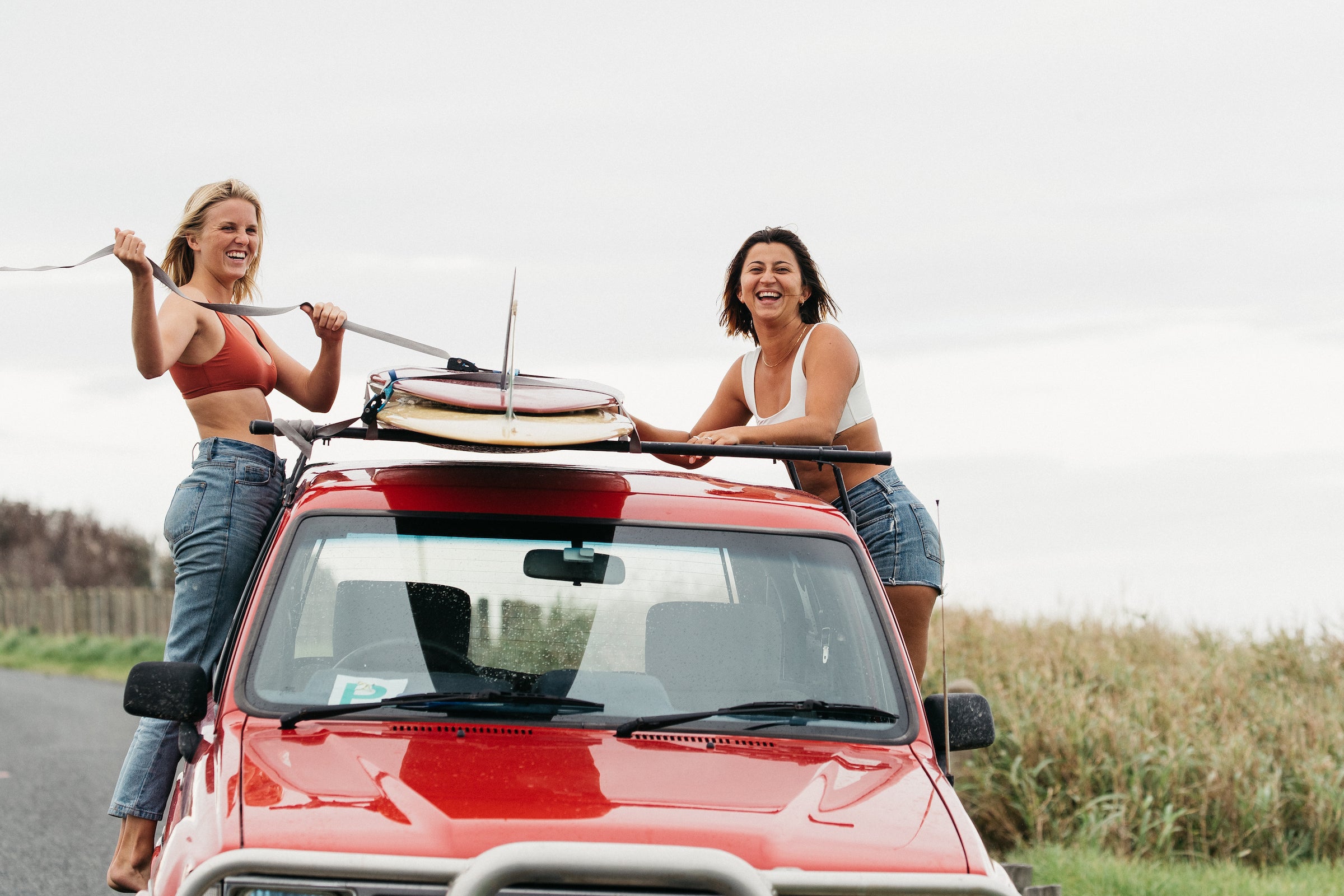 Two woman laughing and strapping down their longboard surf boards to the top of a red toyota truck
