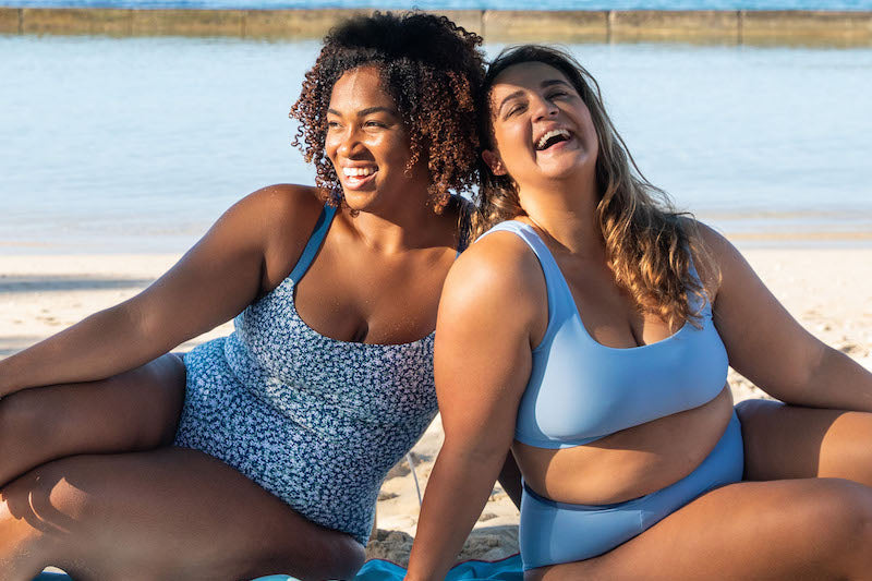 Two women sitting on the beach, one wearing the Paris One Piece in Morning Glory Print, and the other wearing the Jane Surf Bikini top and Alice Surf Bikini bottom in Sky Blue
