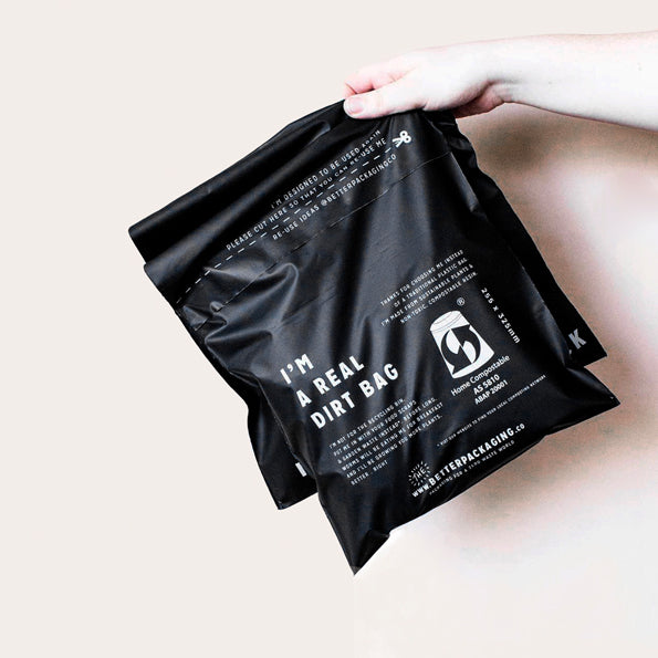 Hand hold two compostable black shipping mailer bags against an ivory wall. 