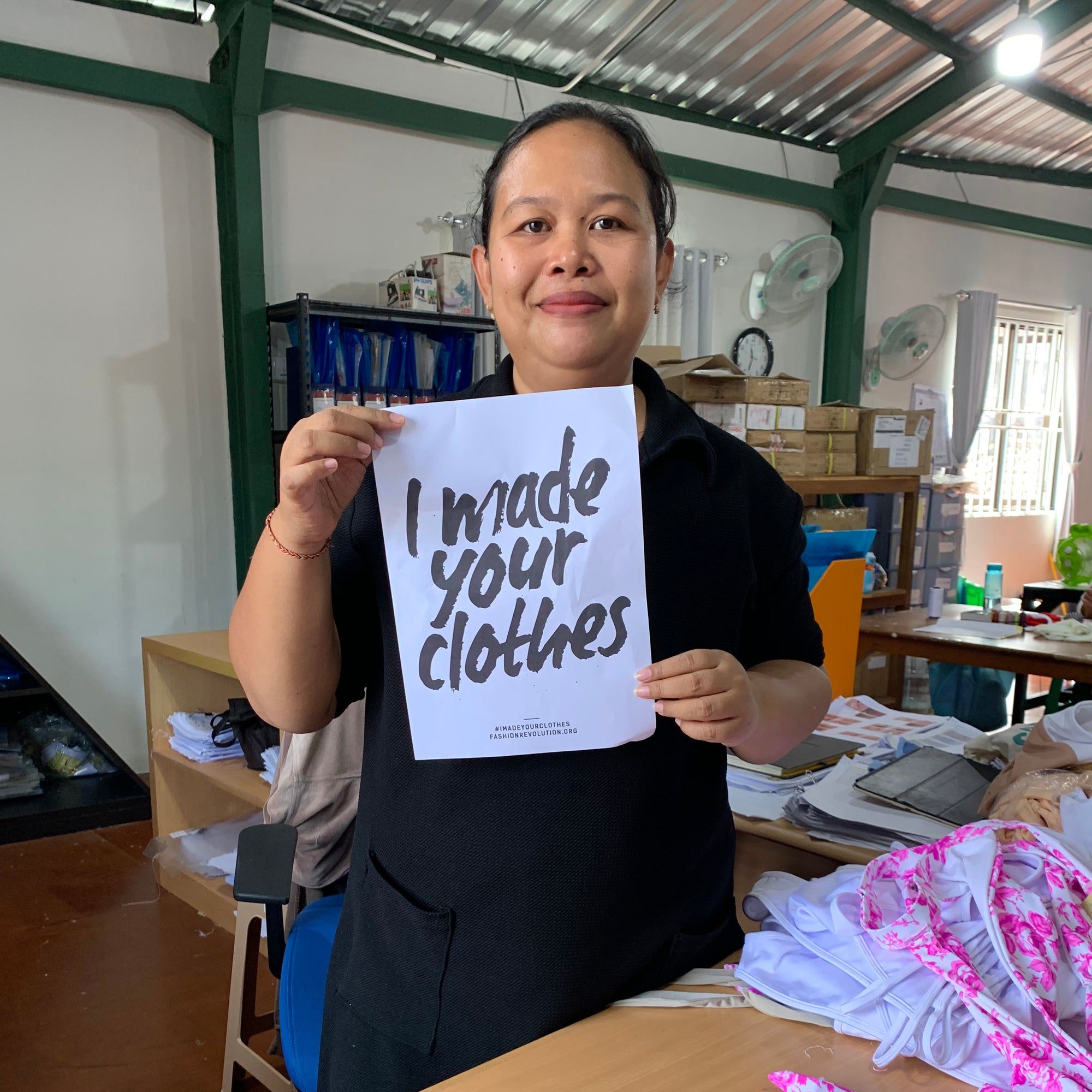 Woman smiling standing in a factory holding a sign that says I made your Cloths
