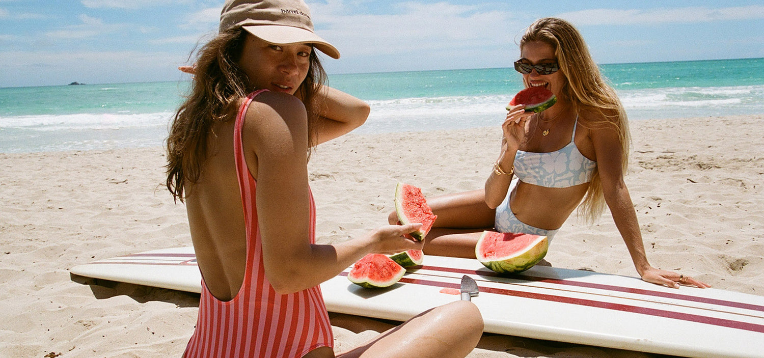 Two women enjoying watermelon on a beach with a surfboard and ocean view.