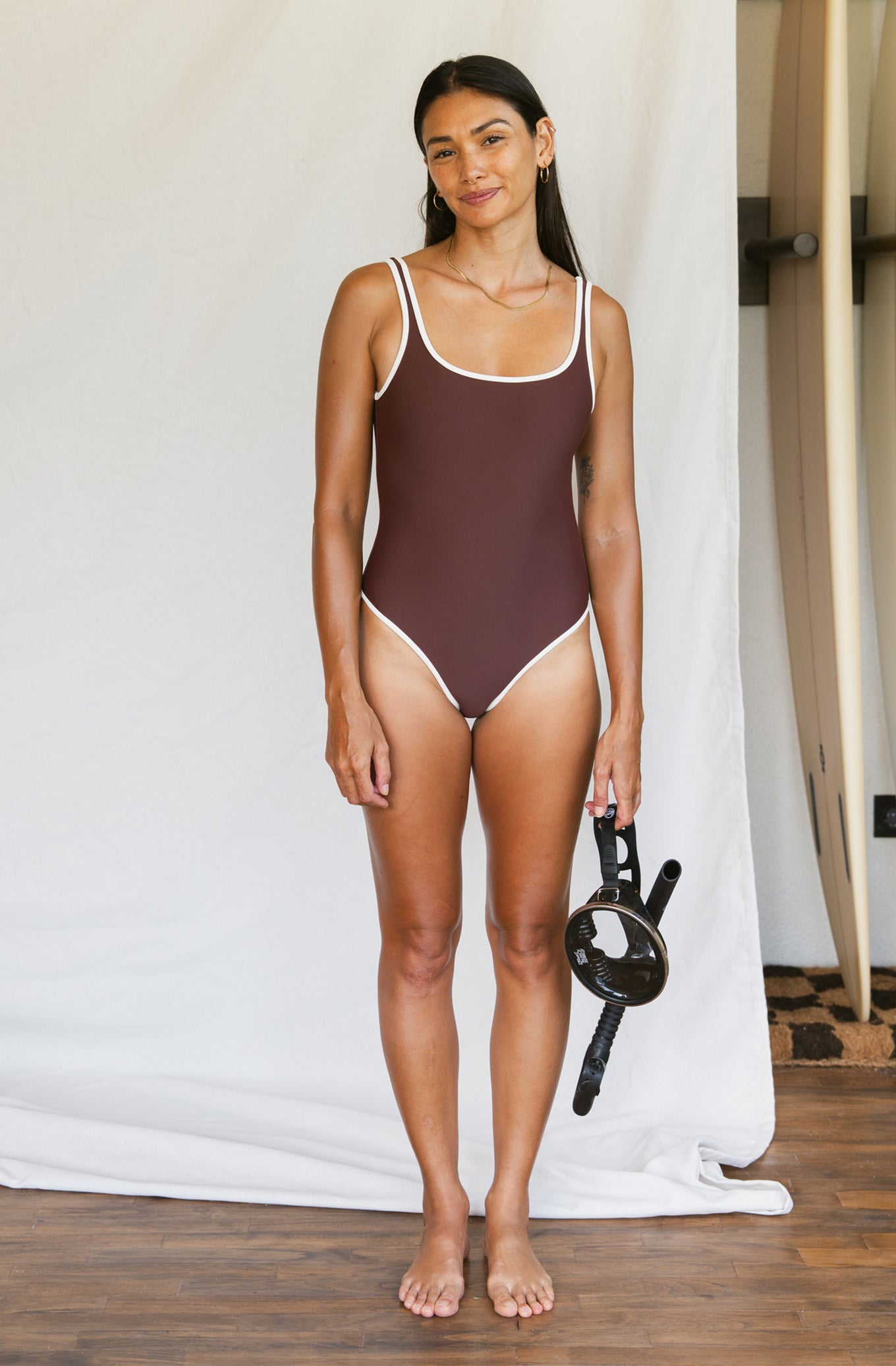 Woman wearing a brown swimsuit standing against a white curtain.