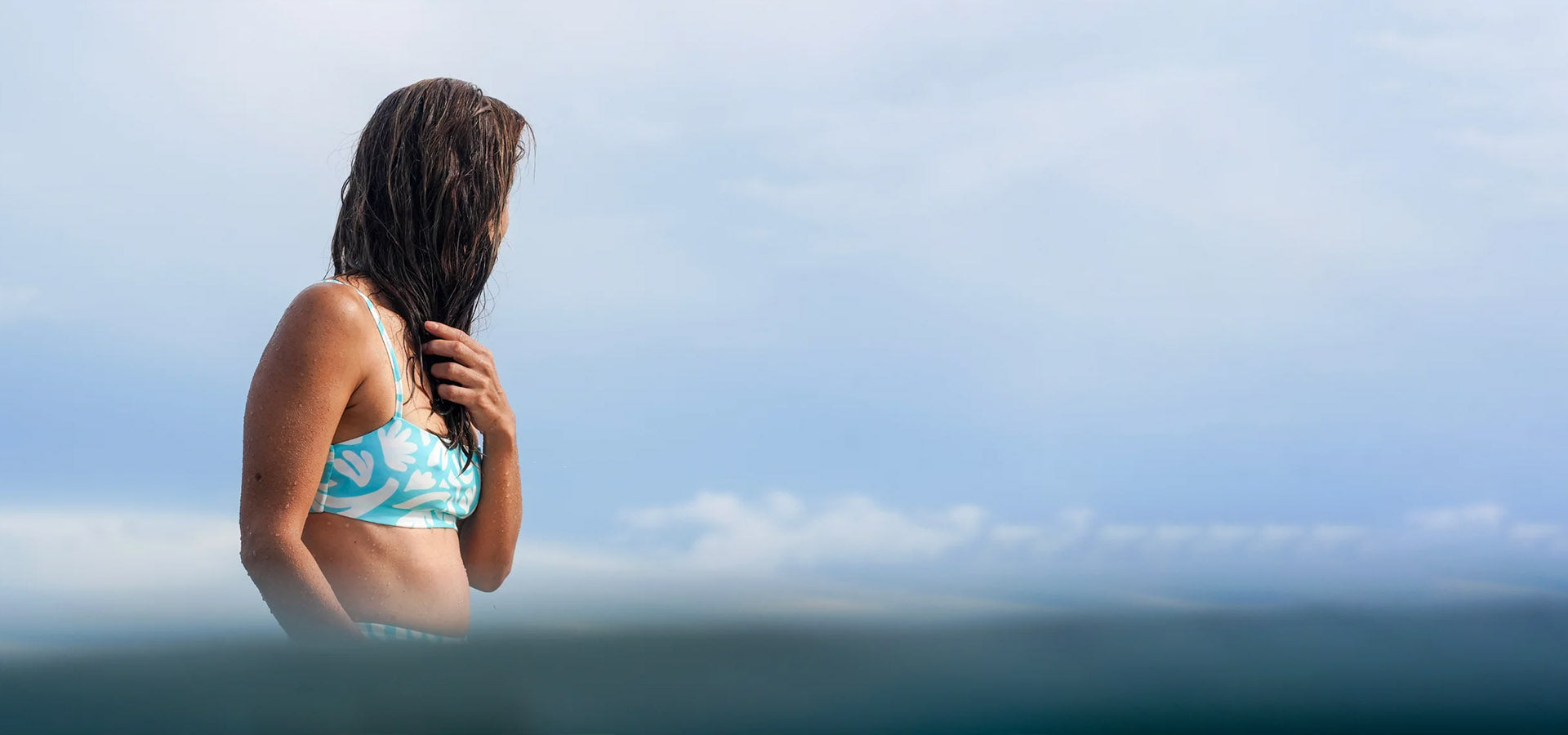 Woman in a bikini standing on a cliff overlooking the ocean with a clear blue sky.