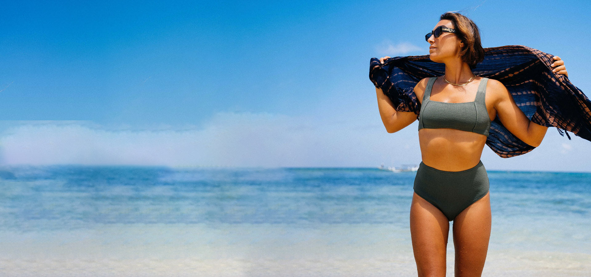 Woman in a green swimsuit standing on a beach with clear blue water and sky.