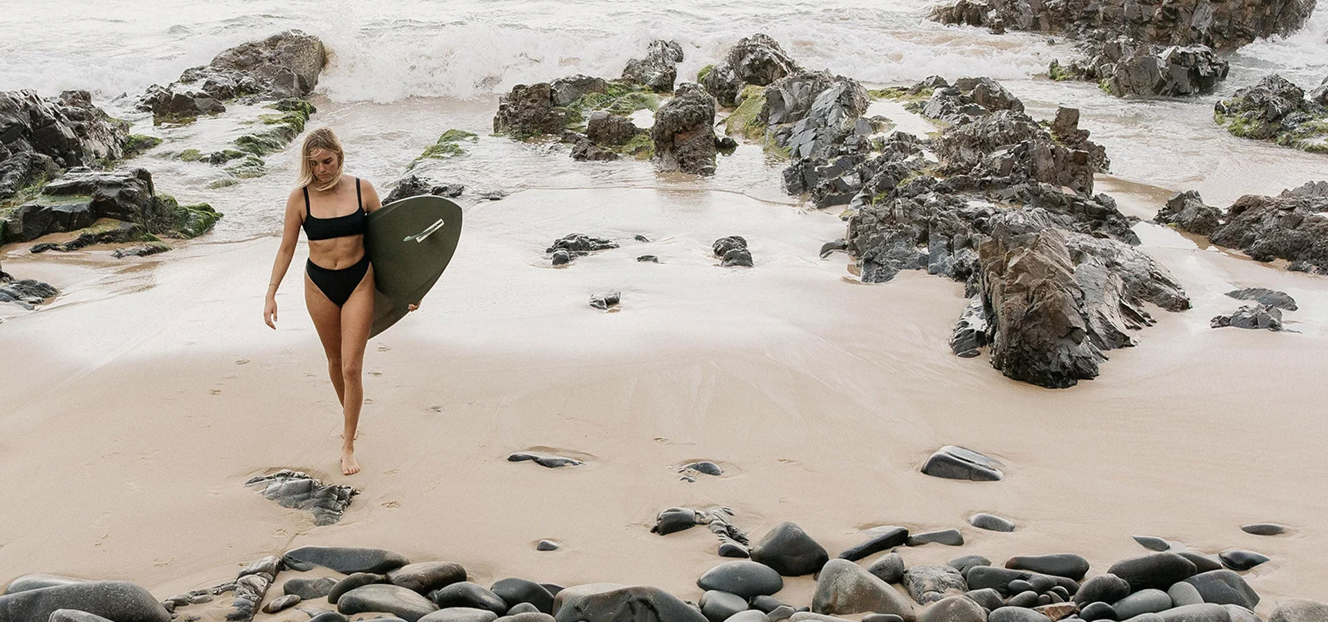 Woman walking on a beach with a surfboard, surrounded by rocks and water.