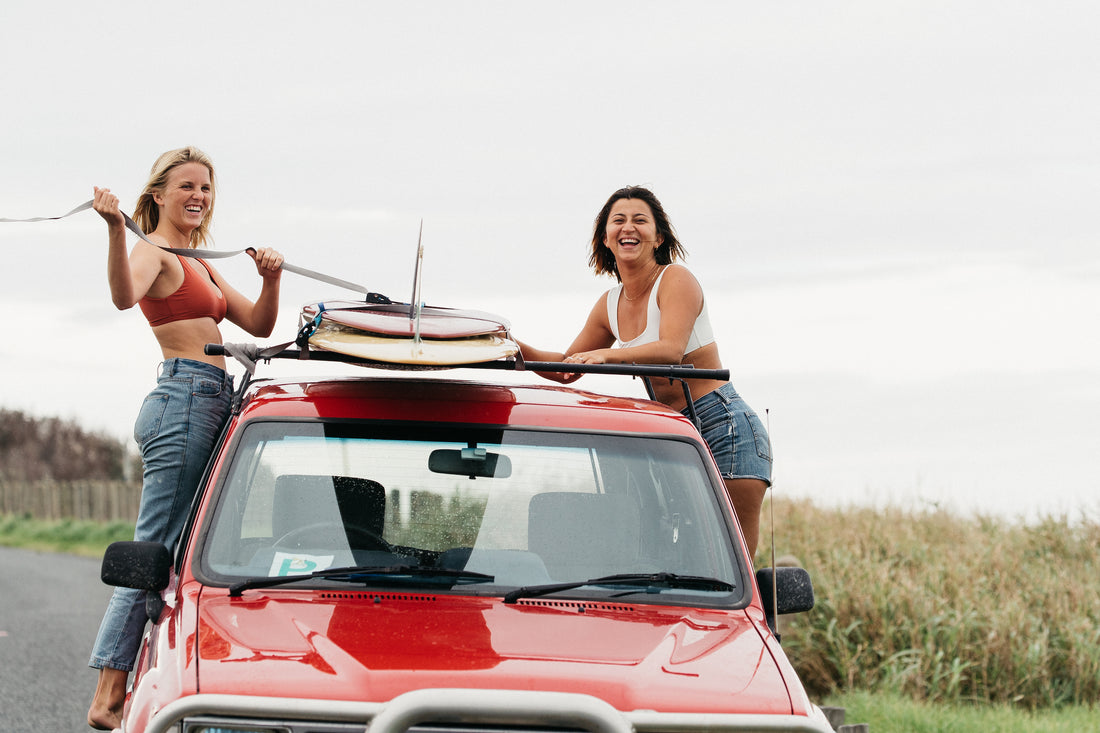 Two woman laughing and strapping down their longboard surf boards to the top of a red toyota truck