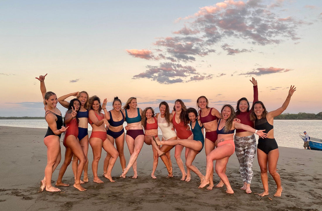 group of 15 women wearing surf bikinis and one piece swimsuits standing on a beach with arms around each other some with their hands in the air 