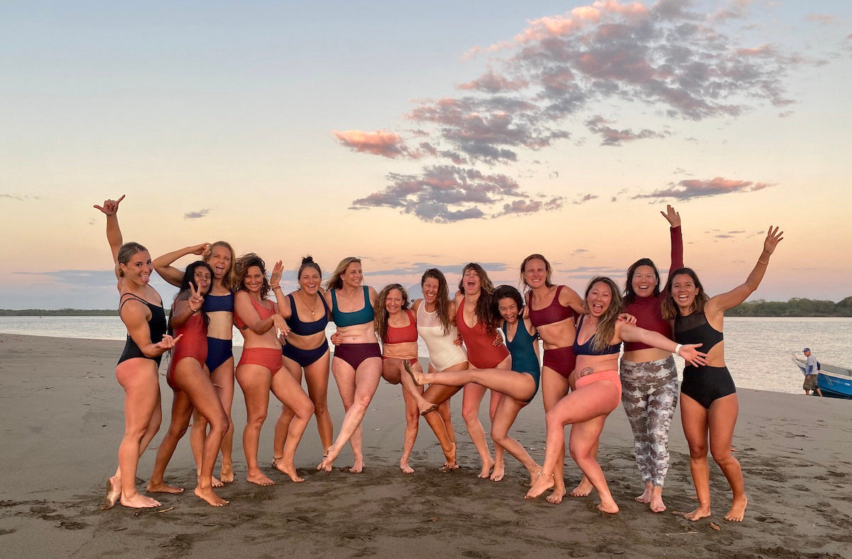 group of 15 women wearing surf bikinis and one piece swimsuits standing on a beach with arms around each other some with their hands in the air 