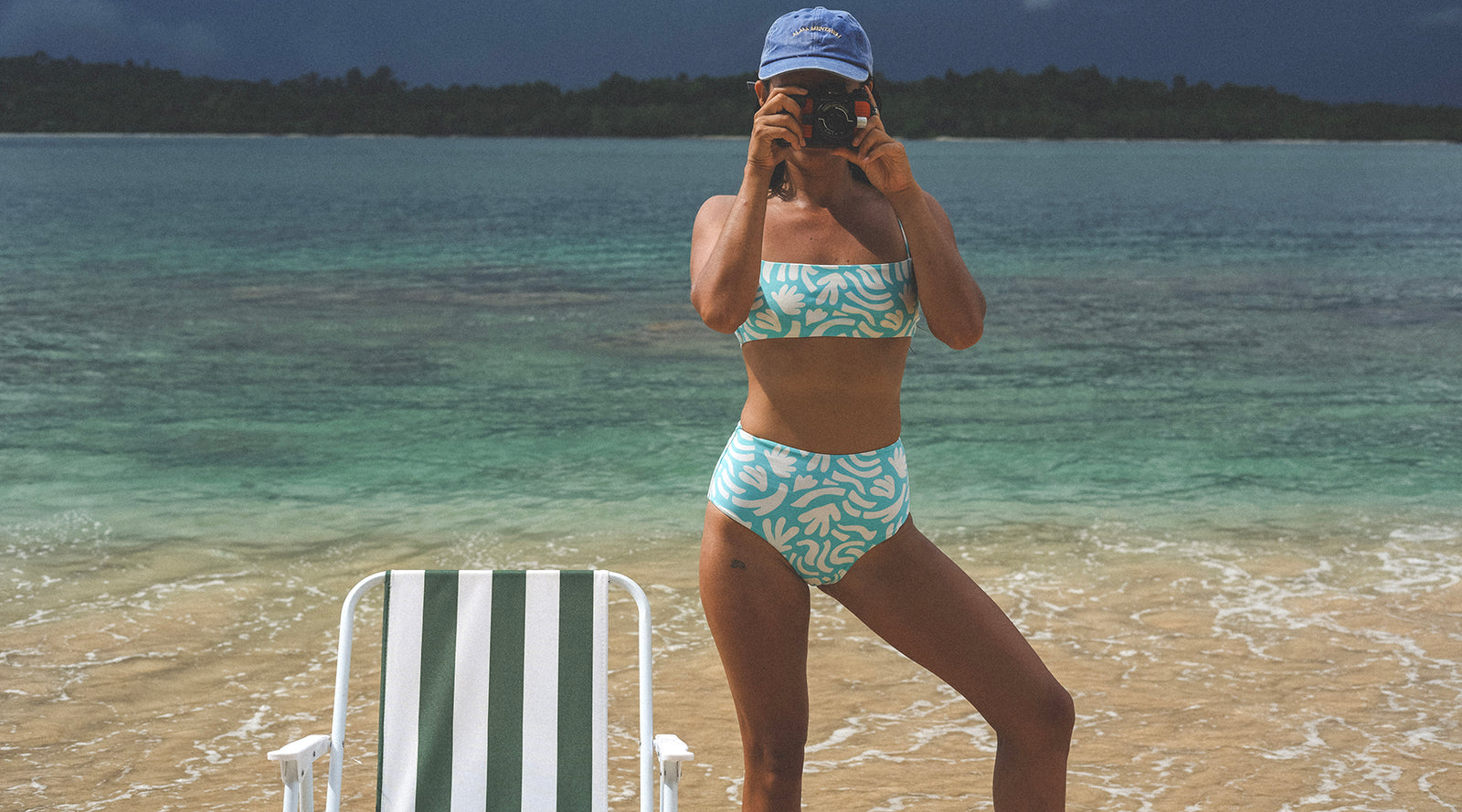 Woman in a swimsuit taking a selfie on a beach with clear water and a chair in the foreground.