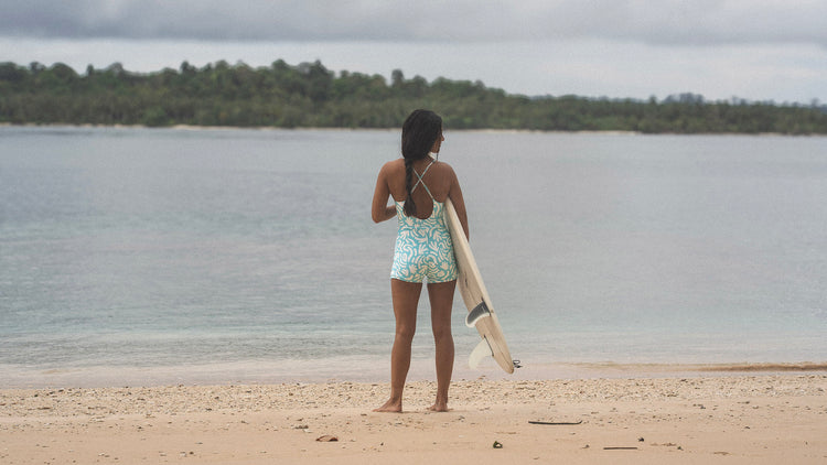 Person standing on a beach holding a surfboard, looking out at the water with trees in the background.