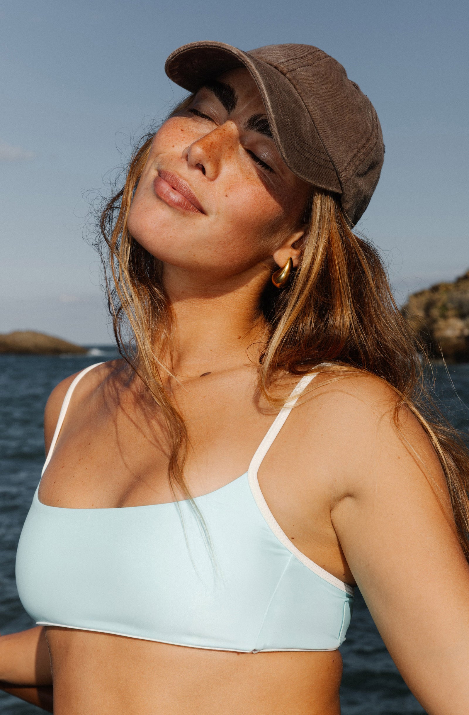 Woman in a light blue bikini standing by a rocky coastline with cliffs and ocean.