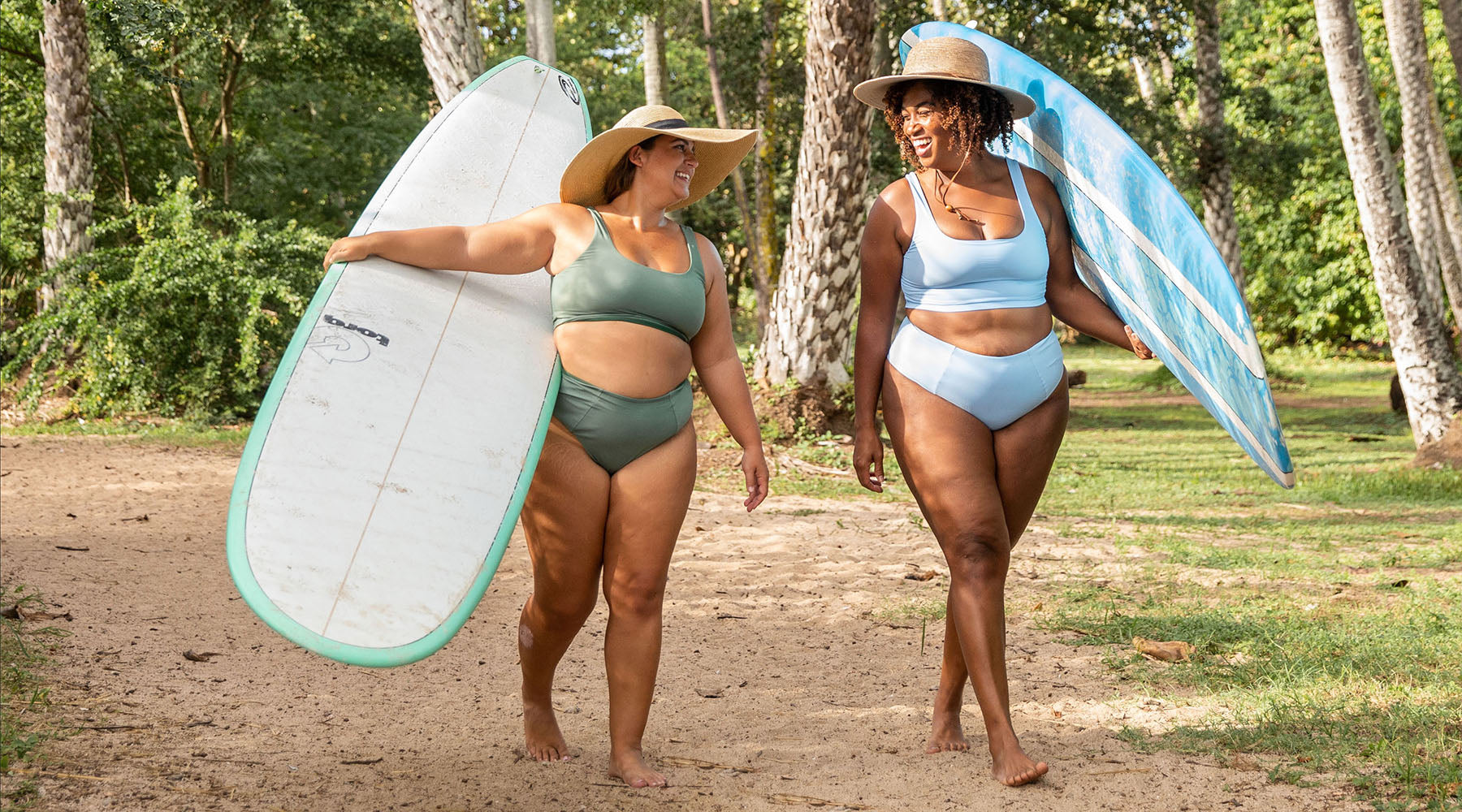 2 women walking on a path carrying surf boards in bikinis