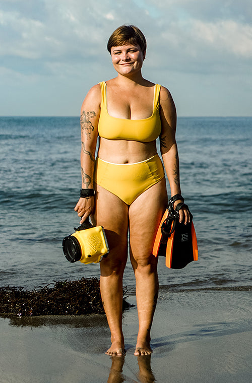 Portrait of woman standing on the beach wearing the Eden surf bikini to and Lucky surf bikini bottom in Yellow holding her water camera and swim fins