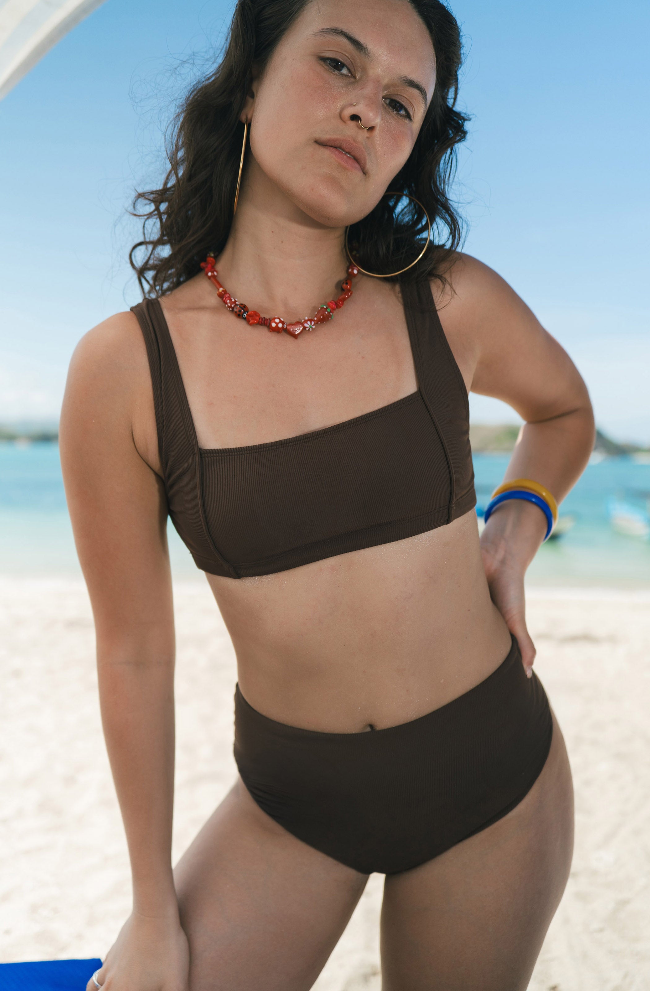 A woman wearing a dark brown two-piece bikini stands on a sandy beach with the ocean and blue sky in the background.