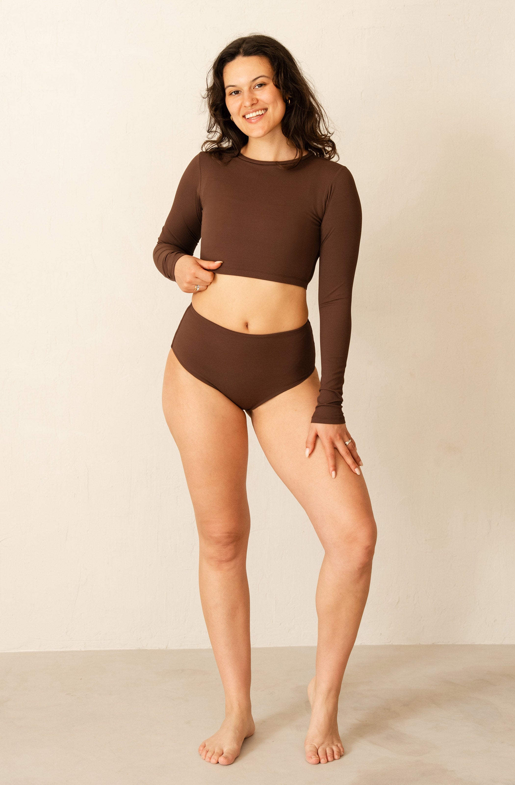 A woman modeling a matching two-piece swimsuit or lounge set in solid dark brown, featuring a long-sleeve cropped top and high-waisted bottoms, standing against a plain beige studio wall.