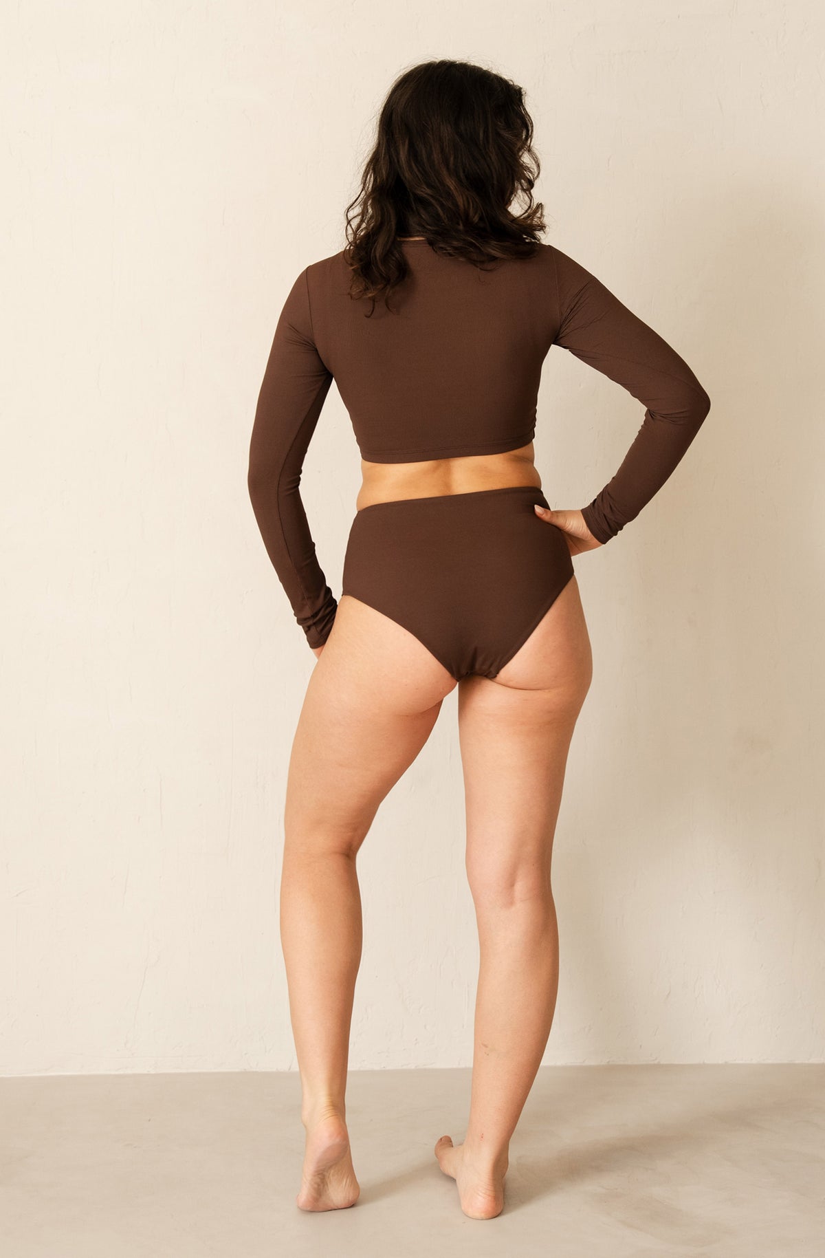 A woman stands with her back to the camera wearing a matching dark brown two-piece swimwear set consisting of a long-sleeve crop top and high-waisted bottoms, set against a plain beige studio background.