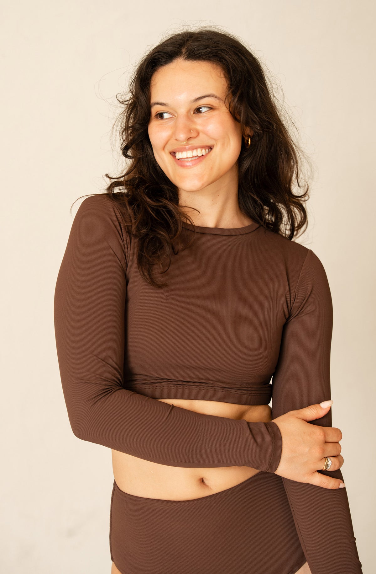A woman with long dark curly hair smiling and looking to the side, wearing a brown long-sleeve crop top and matching high-waisted bottoms against a plain light beige studio background.