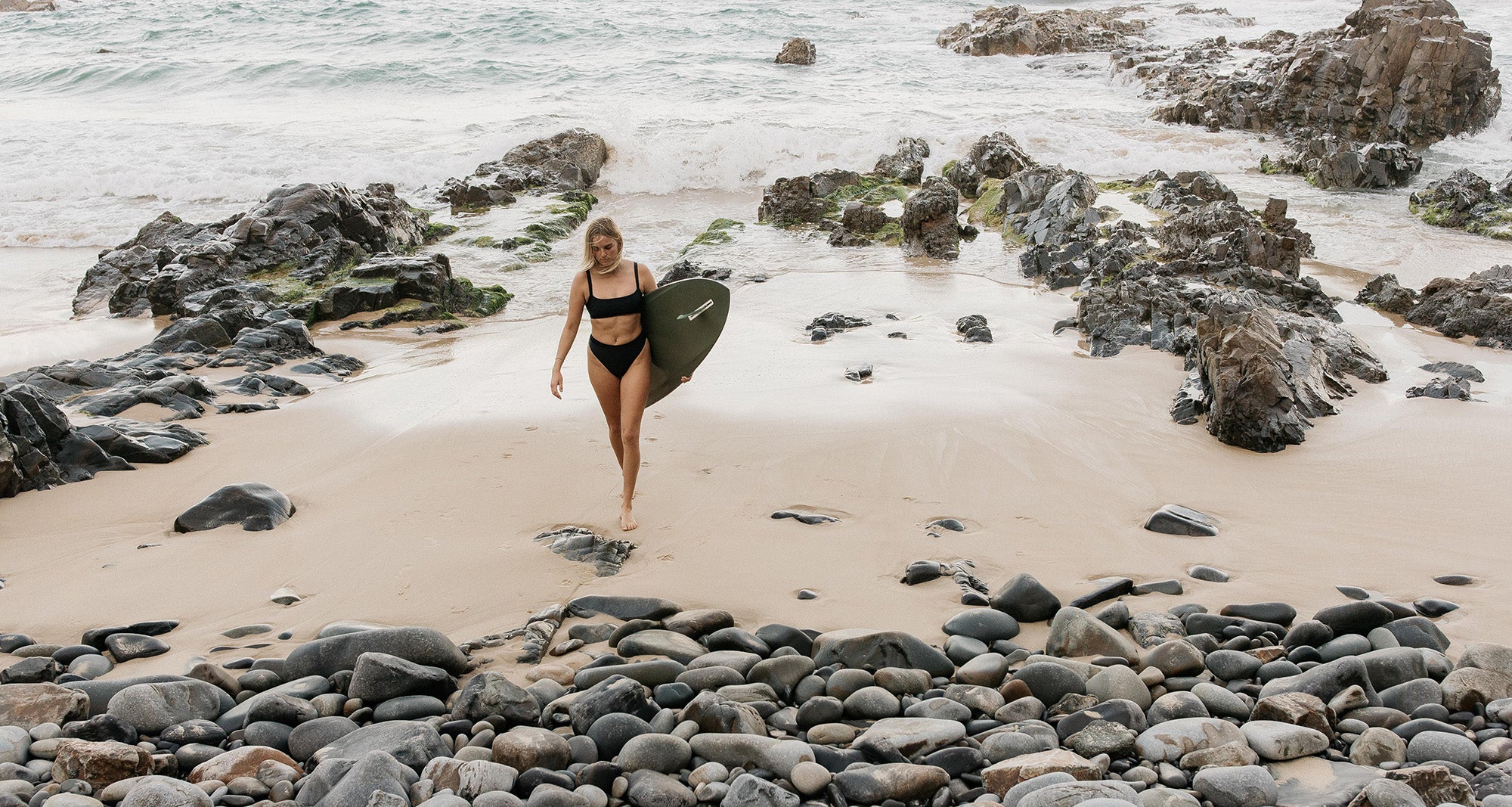 Woman walking out of the water holding a surfboard wearing a black surf bikini on a sandy and rock beach