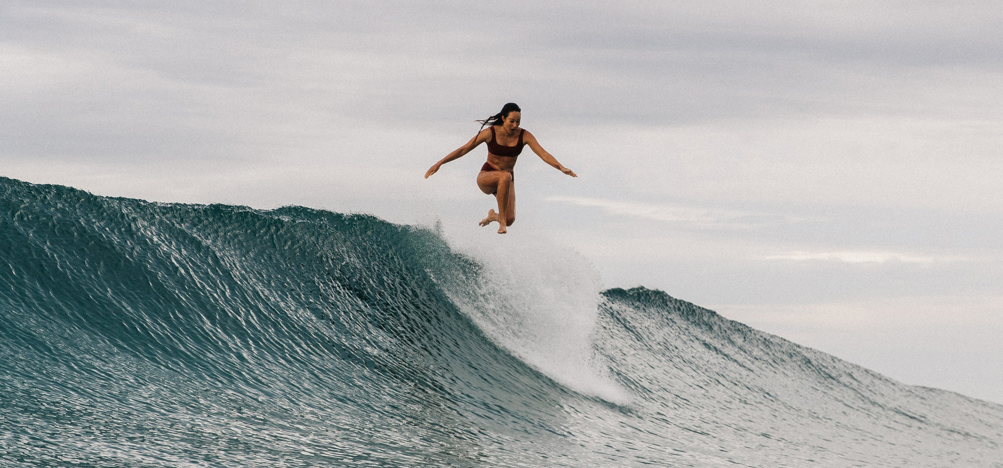 woman jumping off a surfboard in the water wearing a surf bikini top and bottom in rosee