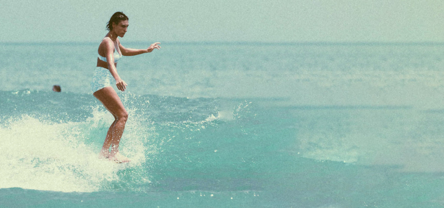 Woman in a bikini standing on a surfboard in clear blue water.