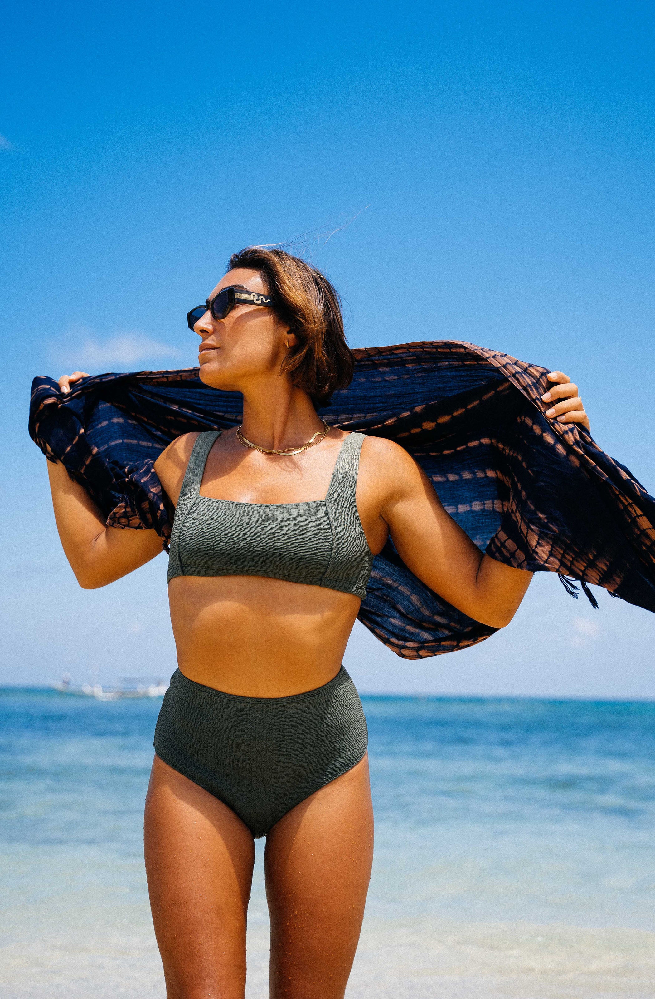 Woman in a green bikini holding a black and blue plaid towel on a beach with clear blue sky and ocean.
