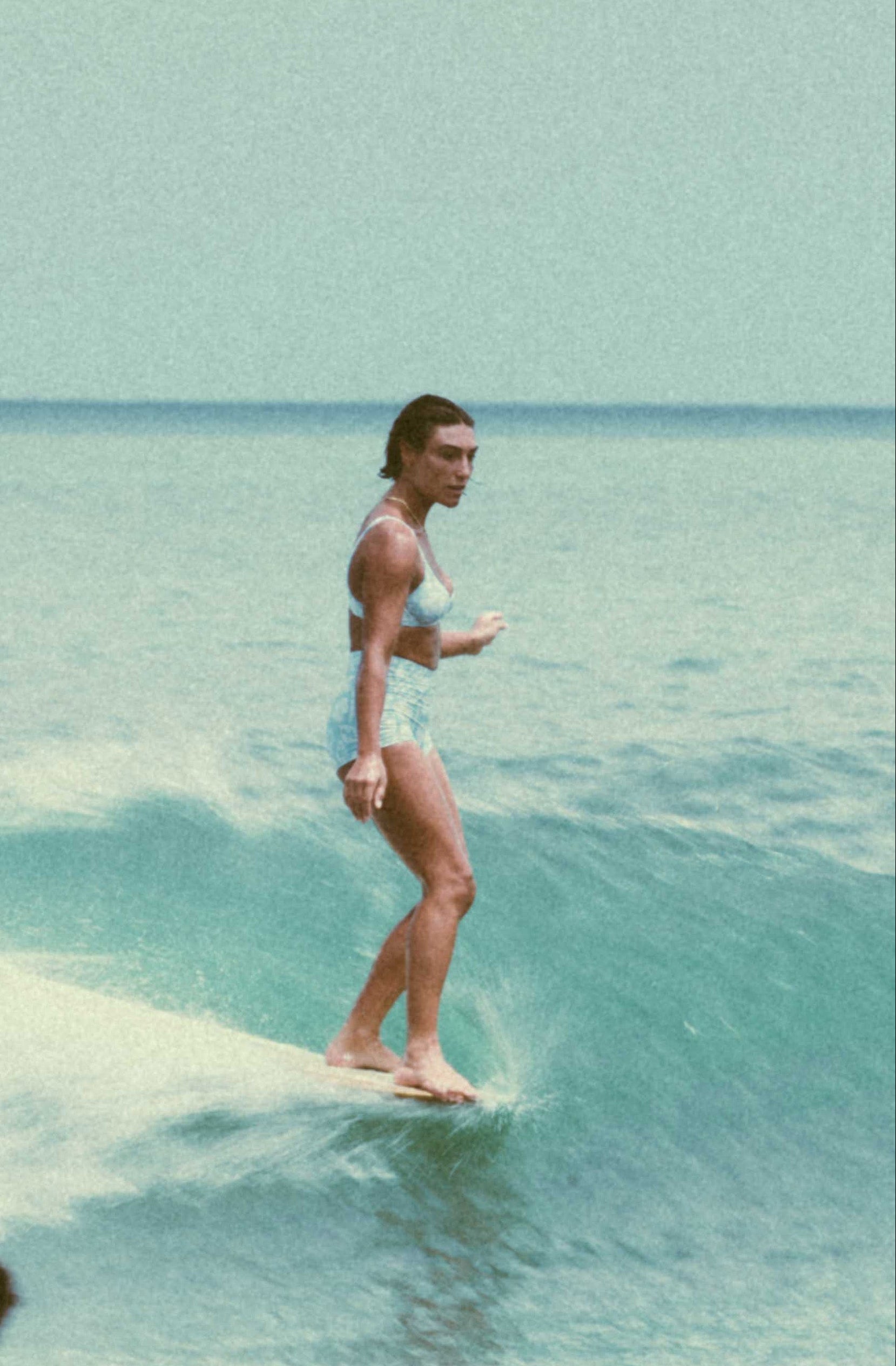 Person surfing on a small wave in clear blue water with another person watching from below.