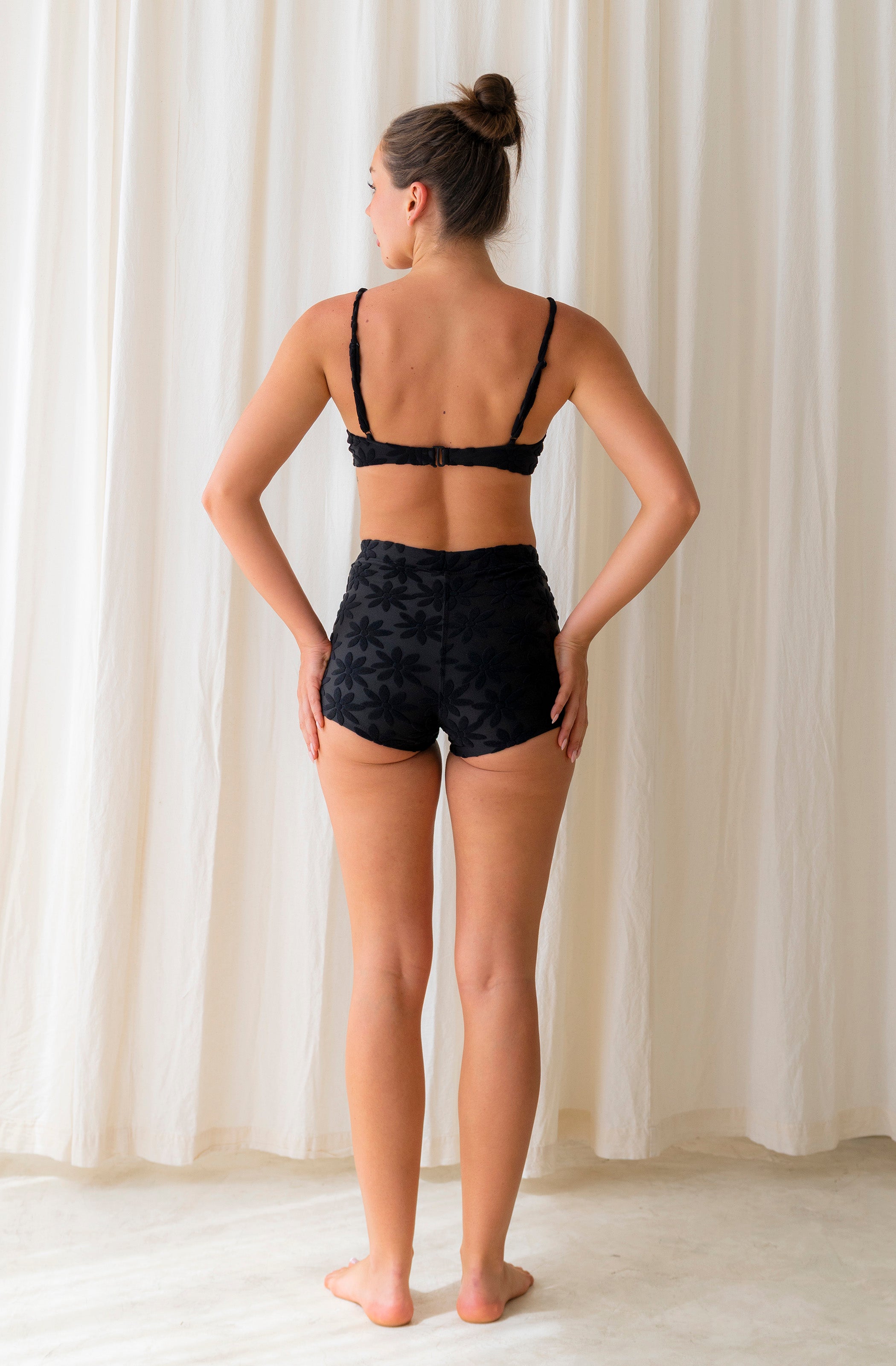 Woman wearing a black bikini top and boyshorts against a white curtain background