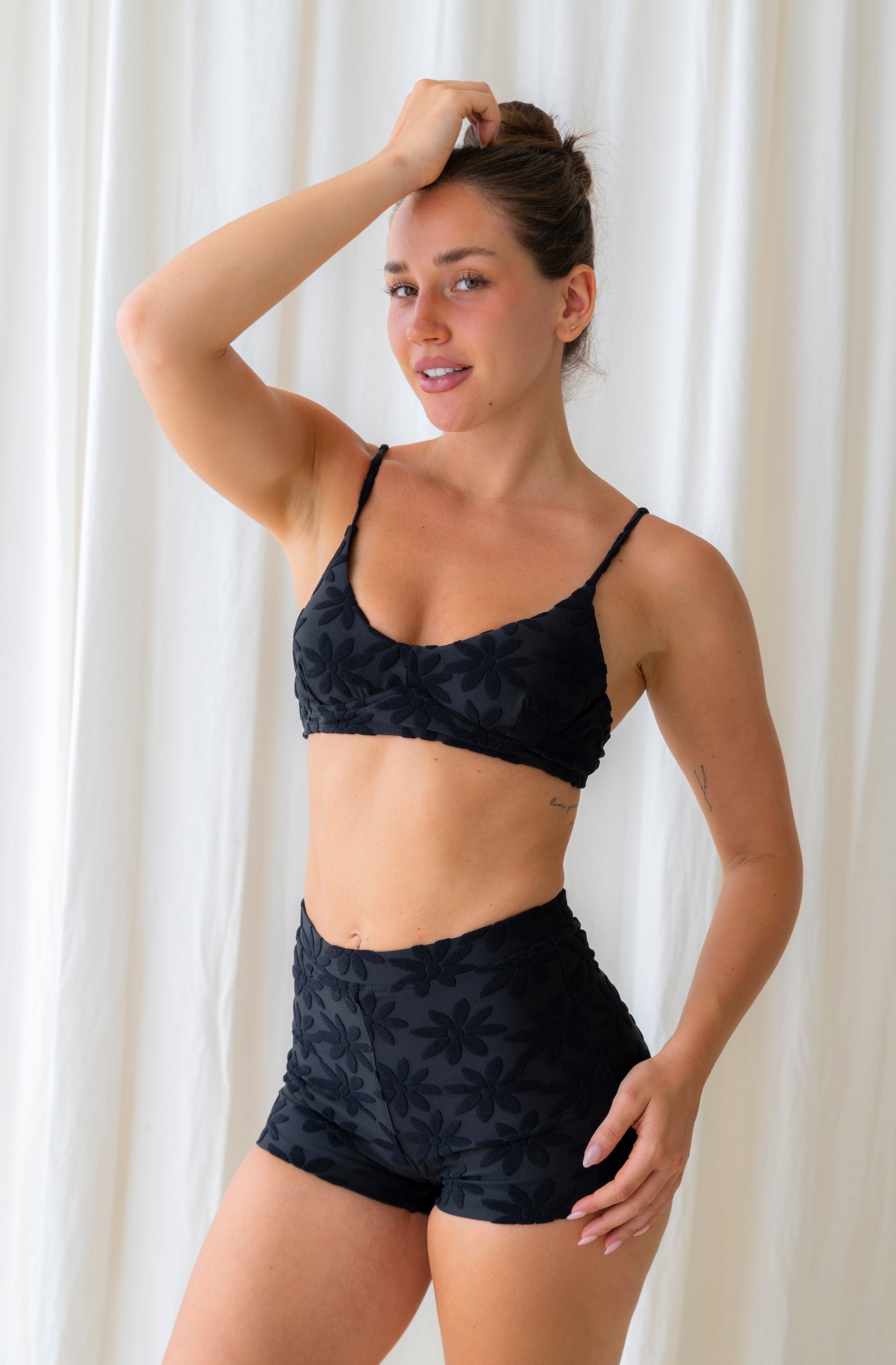 Woman wearing a black bikini top and boyshorts against a white curtain background