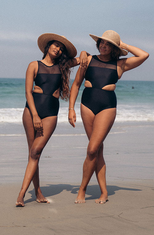 Two woman leaning on each other standing at the beach wearing straw hats and black Malibu One Piece surf swimsuit