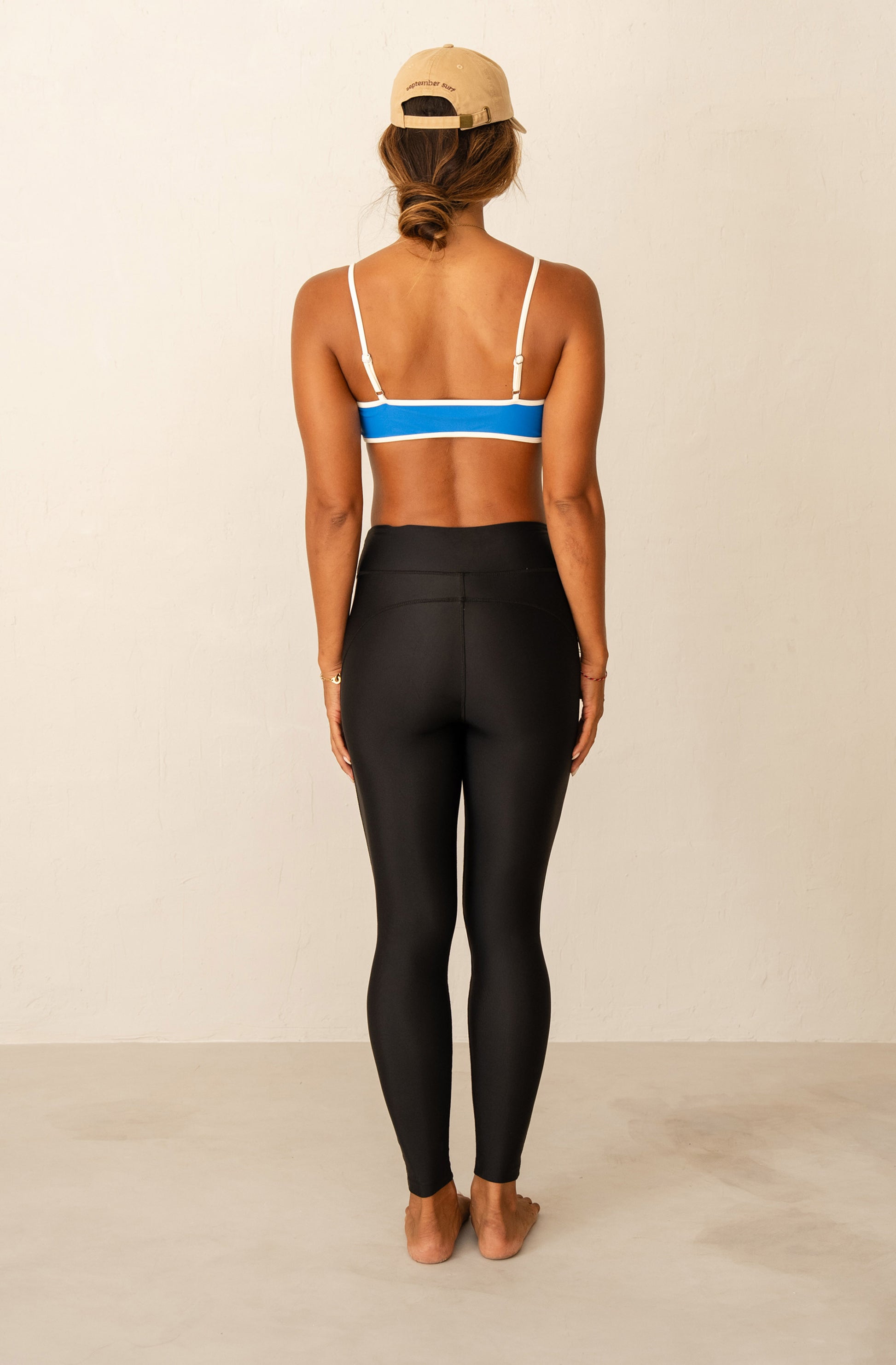 Woman wearing a blue bikini top and black leggings with a beige cap on a light background