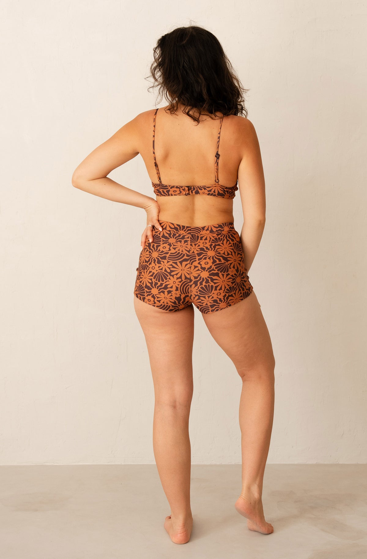 A woman is shown from behind wearing a two-piece bikini set with a brown and black tropical floral pattern, standing against a plain off-white wall.