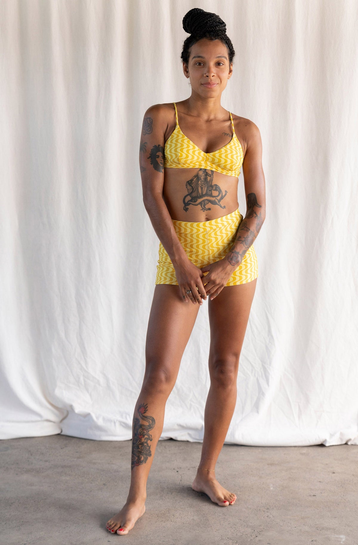A woman modeling a yellow and white patterned two-piece bikini set, consisting of a triangle bikini top and matching high-waisted shorts, standing against a draped white fabric backdrop.