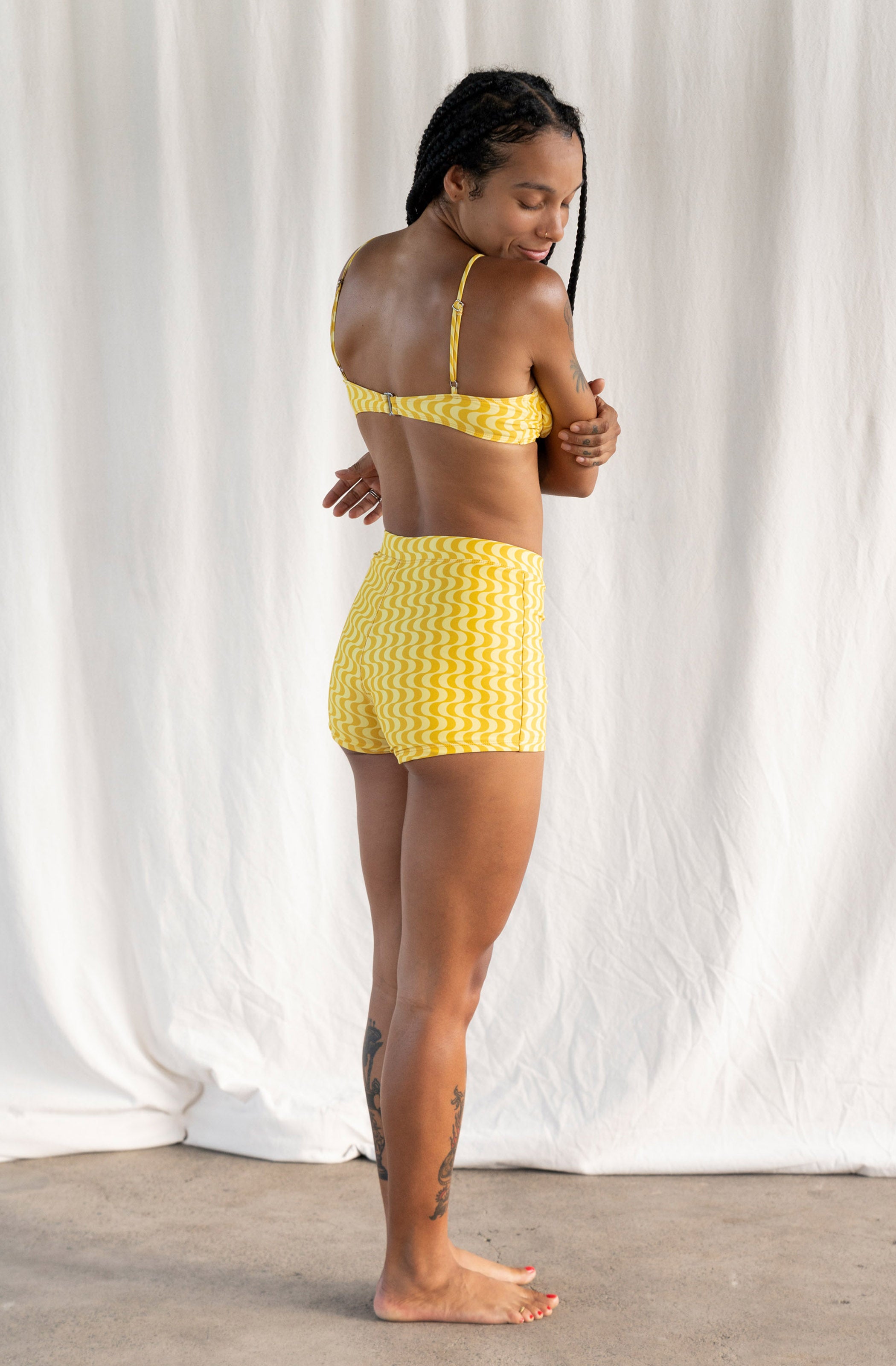 A woman modeling a yellow and white patterned two-piece swimsuit, shown from the back against a white fabric backdrop.