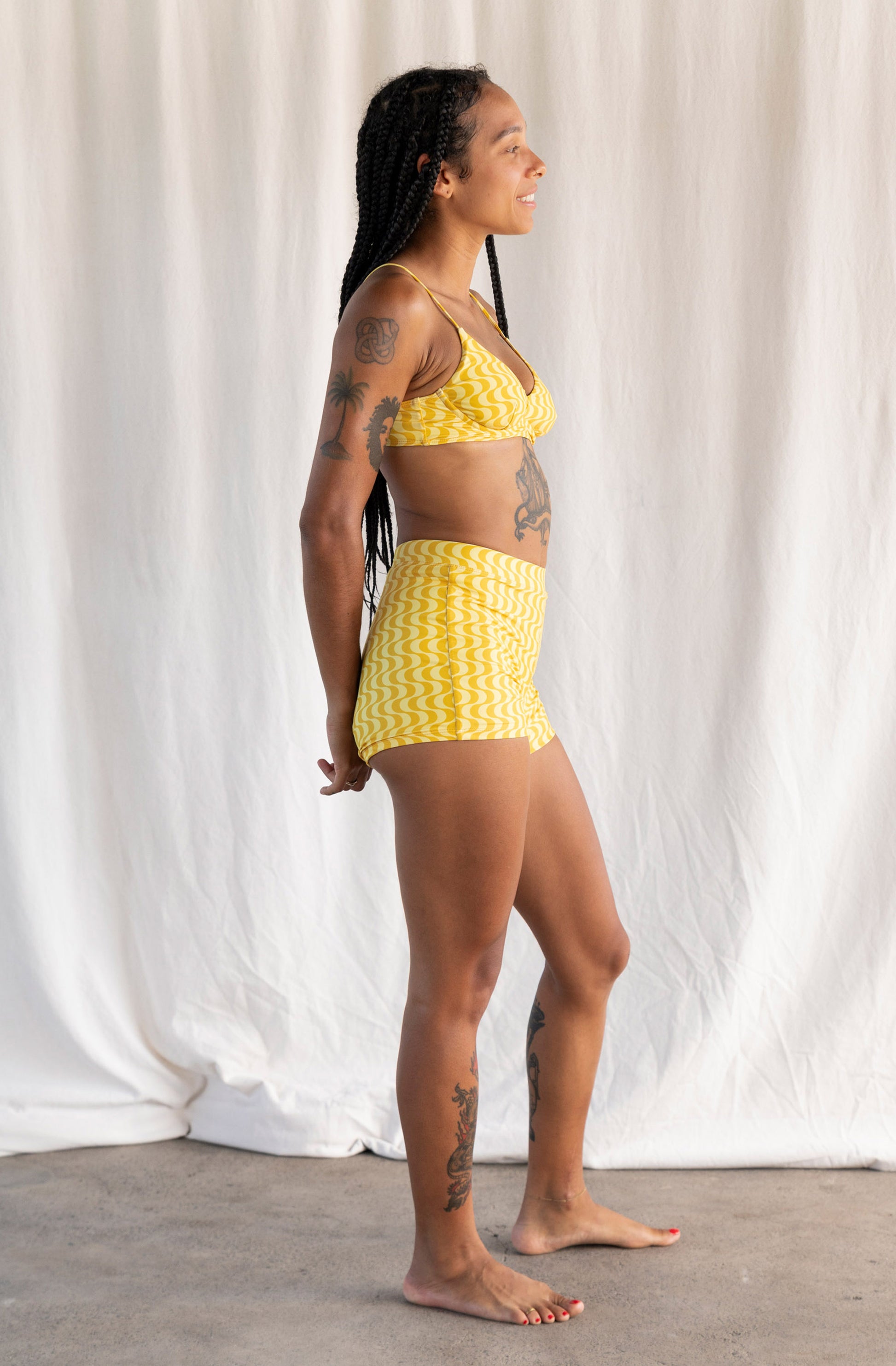 A woman modeling a two-piece bikini and shorts set with a yellow and white chevron pattern, standing in profile against a white fabric backdrop.