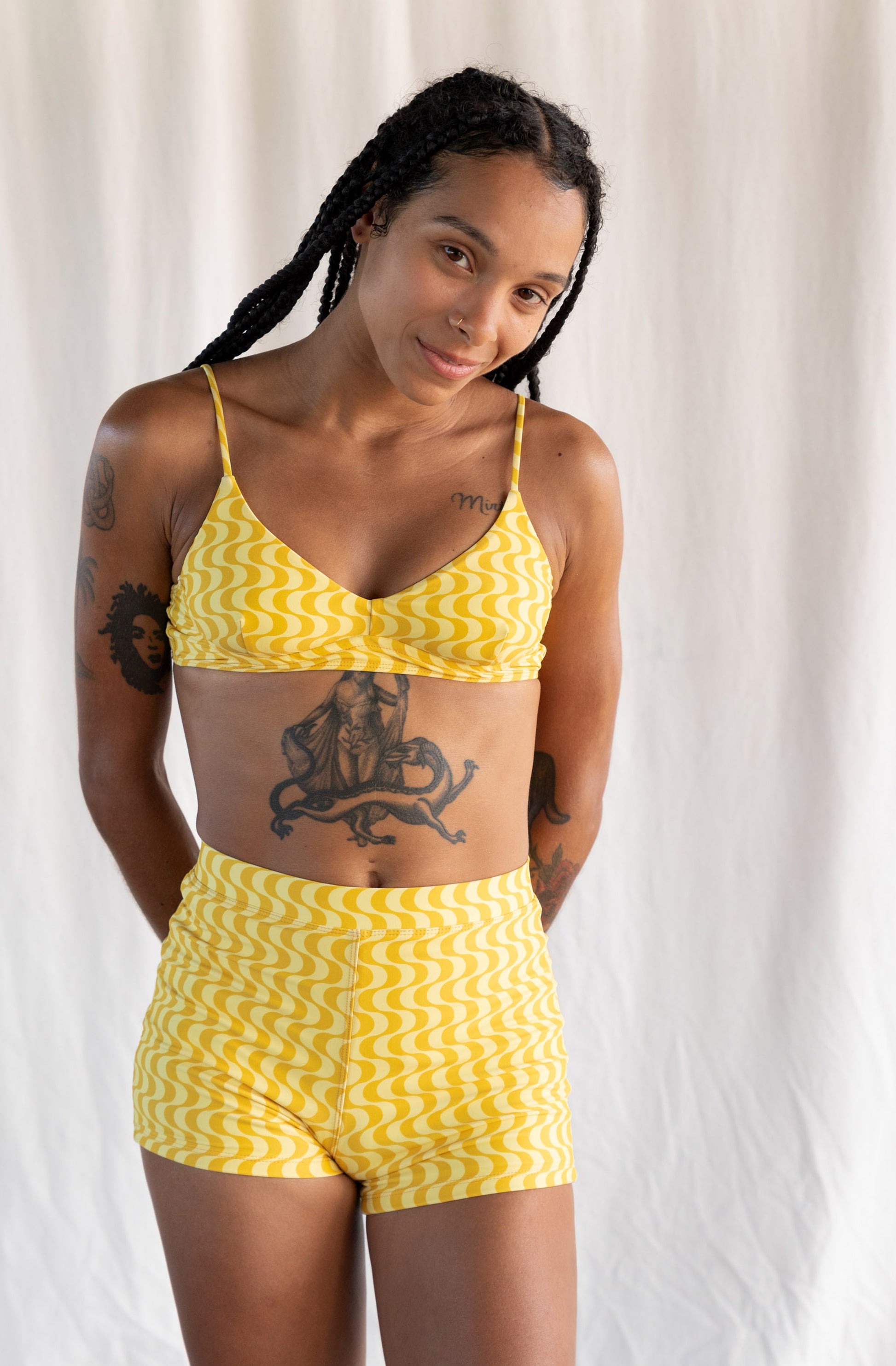 A woman modeling a yellow and white zigzag-patterned two-piece swimsuit, consisting of a triangle bikini top and matching high-waisted shorts, set against a draped white fabric background.