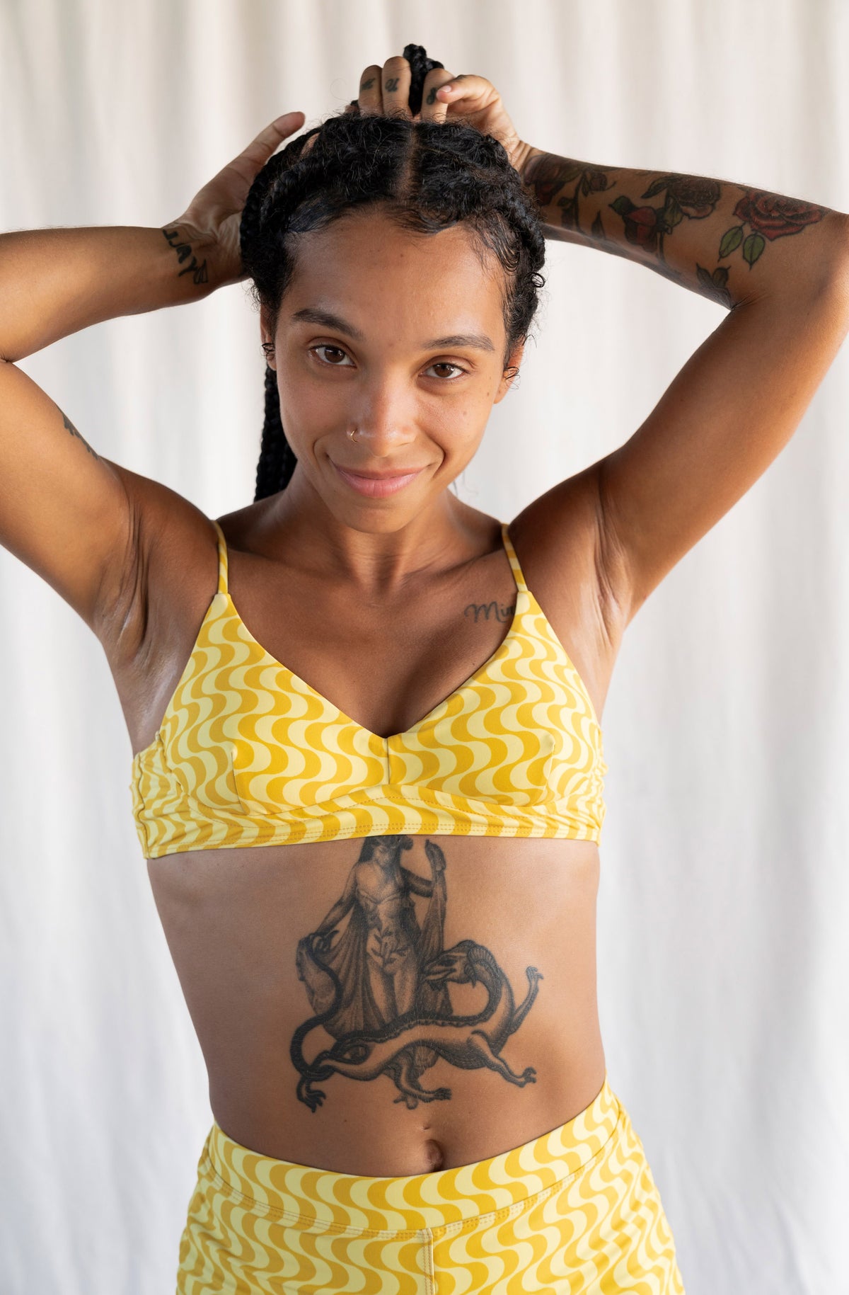 A woman with tattoos modeling a yellow and white wavy patterned two-piece bikini set against a white curtain background.