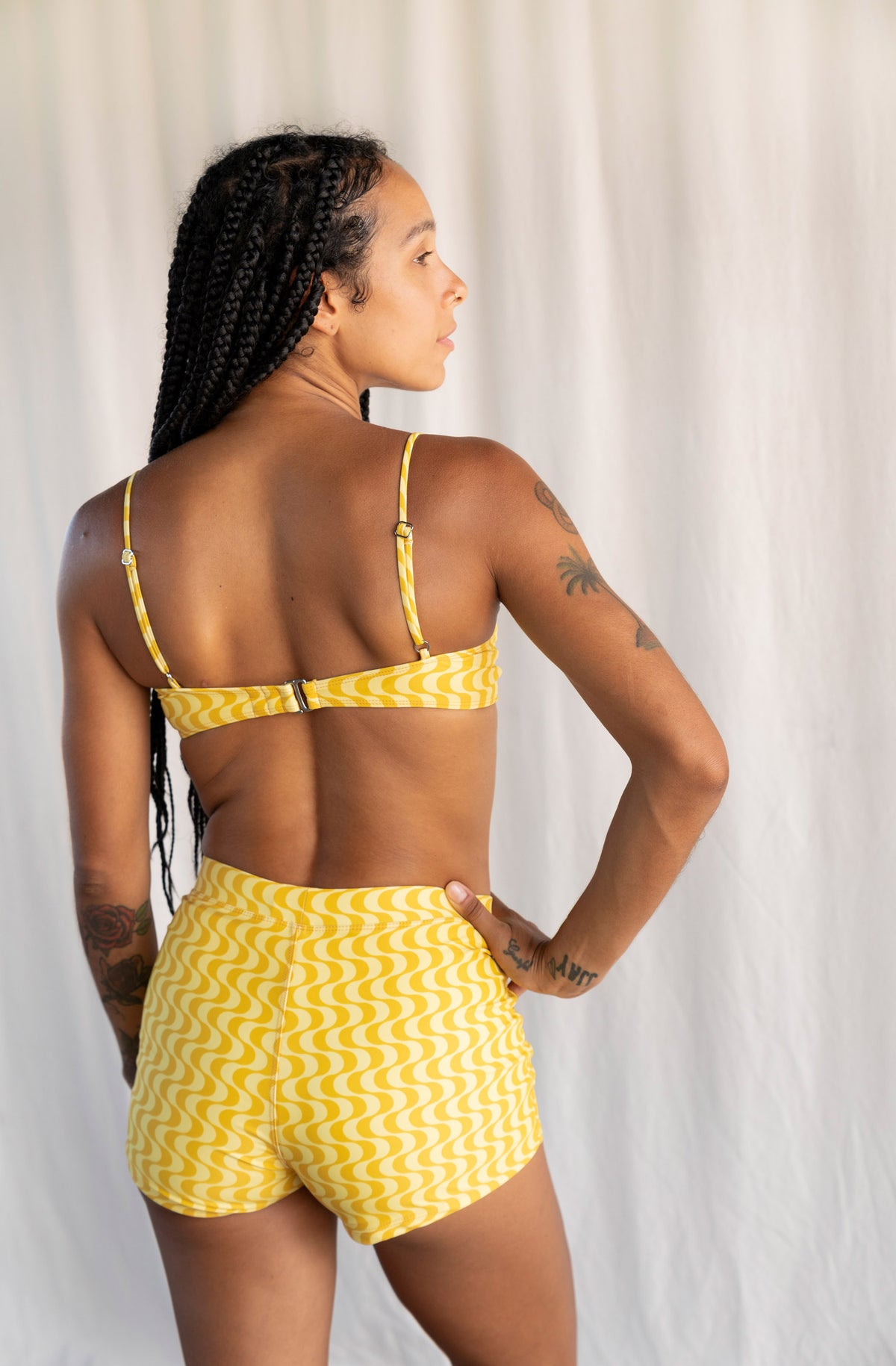 A woman shown from the back wearing a yellow and white wavy-patterned bikini top and matching shorts, standing against a light neutral curtain backdrop.