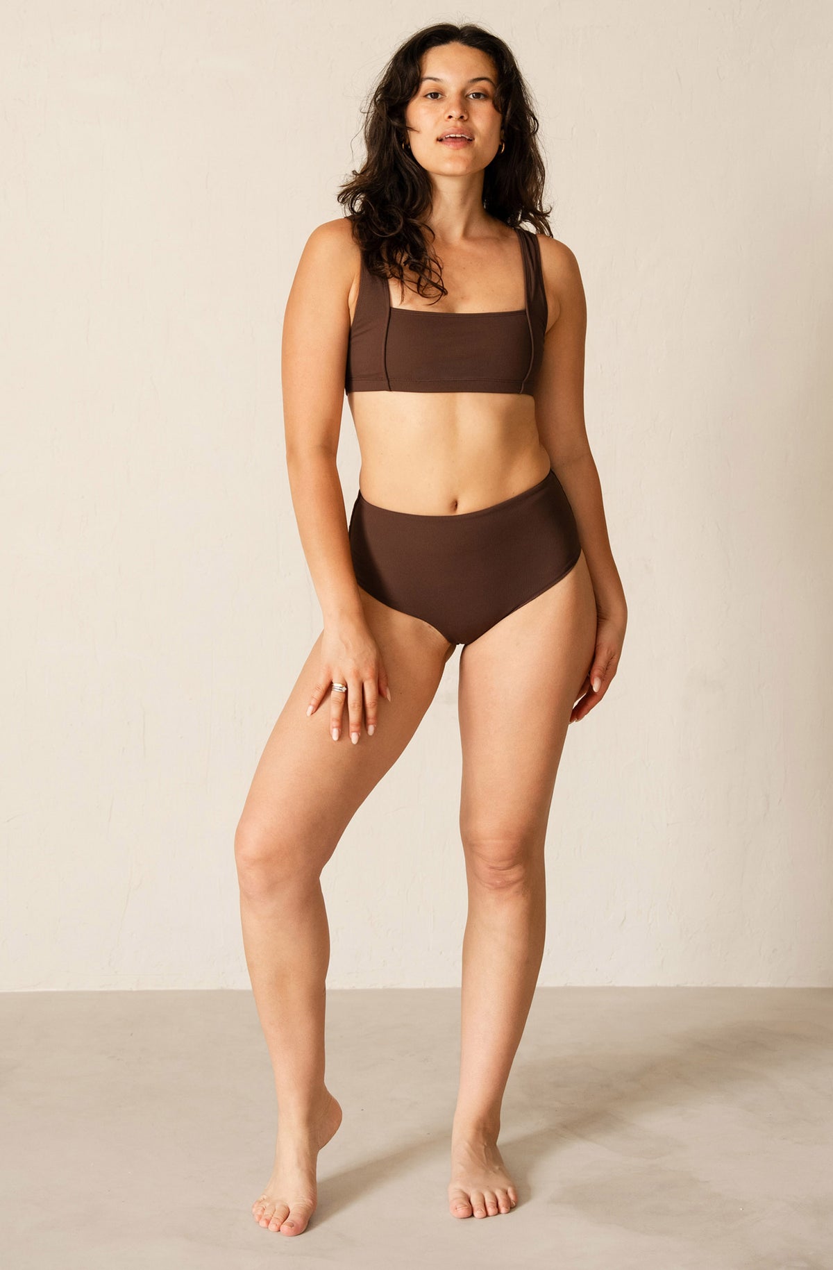 A woman modeling a two-piece brown bikini set, consisting of a square-neck bikini top and high-waisted bikini bottoms, standing against a neutral beige studio background.