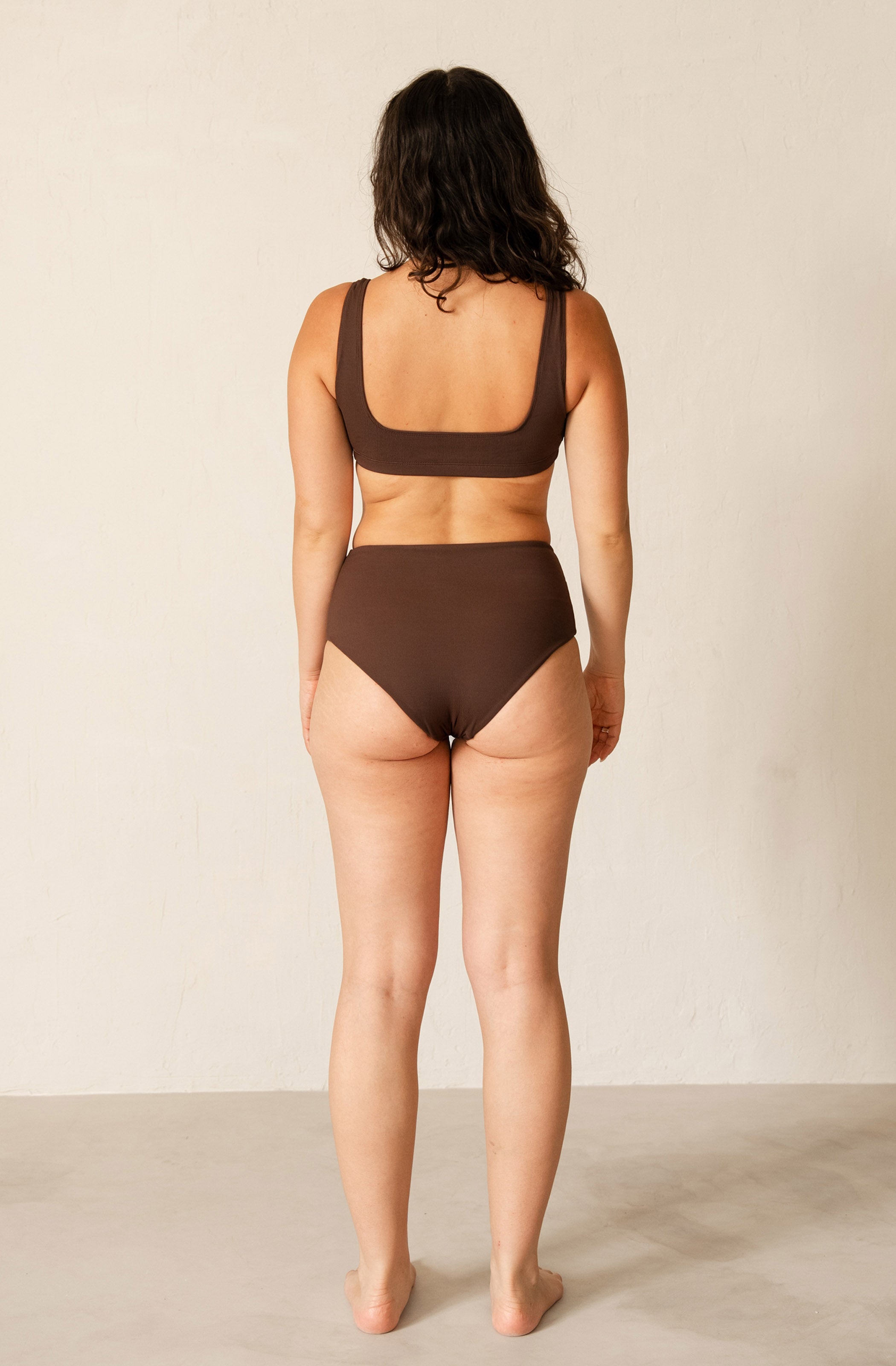 A woman stands with her back to the camera wearing a dark brown two-piece swimsuit, consisting of a scoop-neck bikini top and high-waisted bikini bottoms, against a plain off-white studio background.