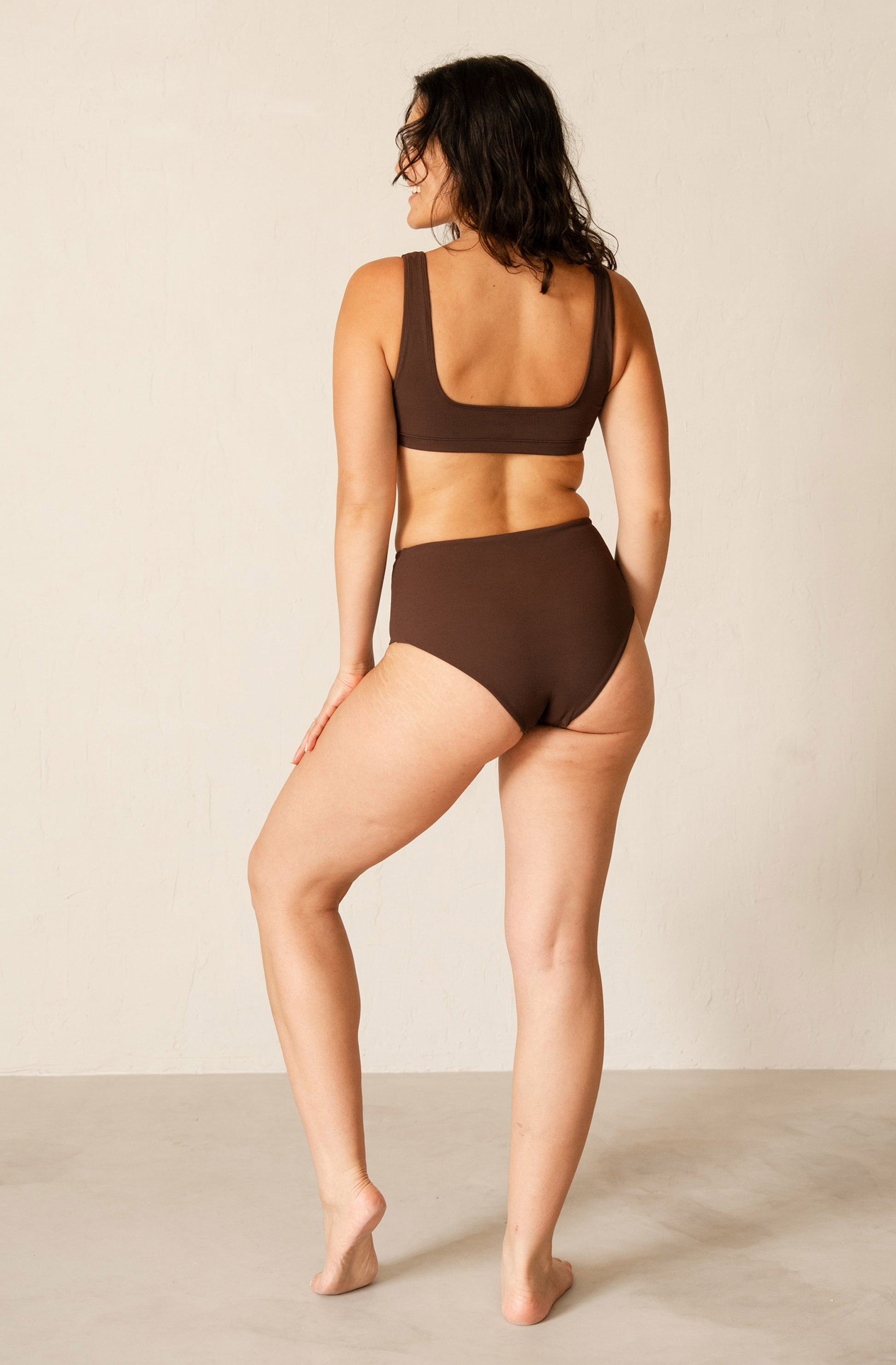 A woman is shown from behind wearing a two-piece brown swimwear set, consisting of a scoop-neck bikini top and high-waisted bikini bottoms, against a neutral beige studio background.