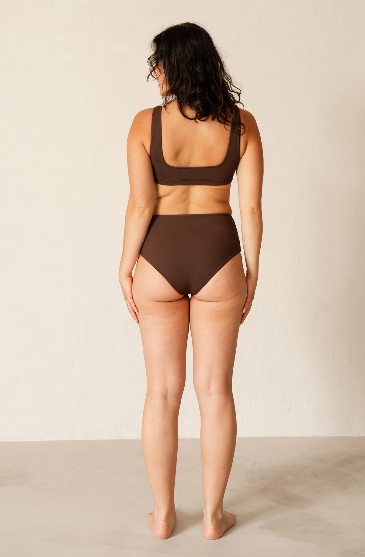 A woman stands with her back to the camera wearing a solid dark brown two-piece swimsuit, featuring a scoop-neck bikini top and high-waisted bikini bottoms, set against a plain off-white wall.