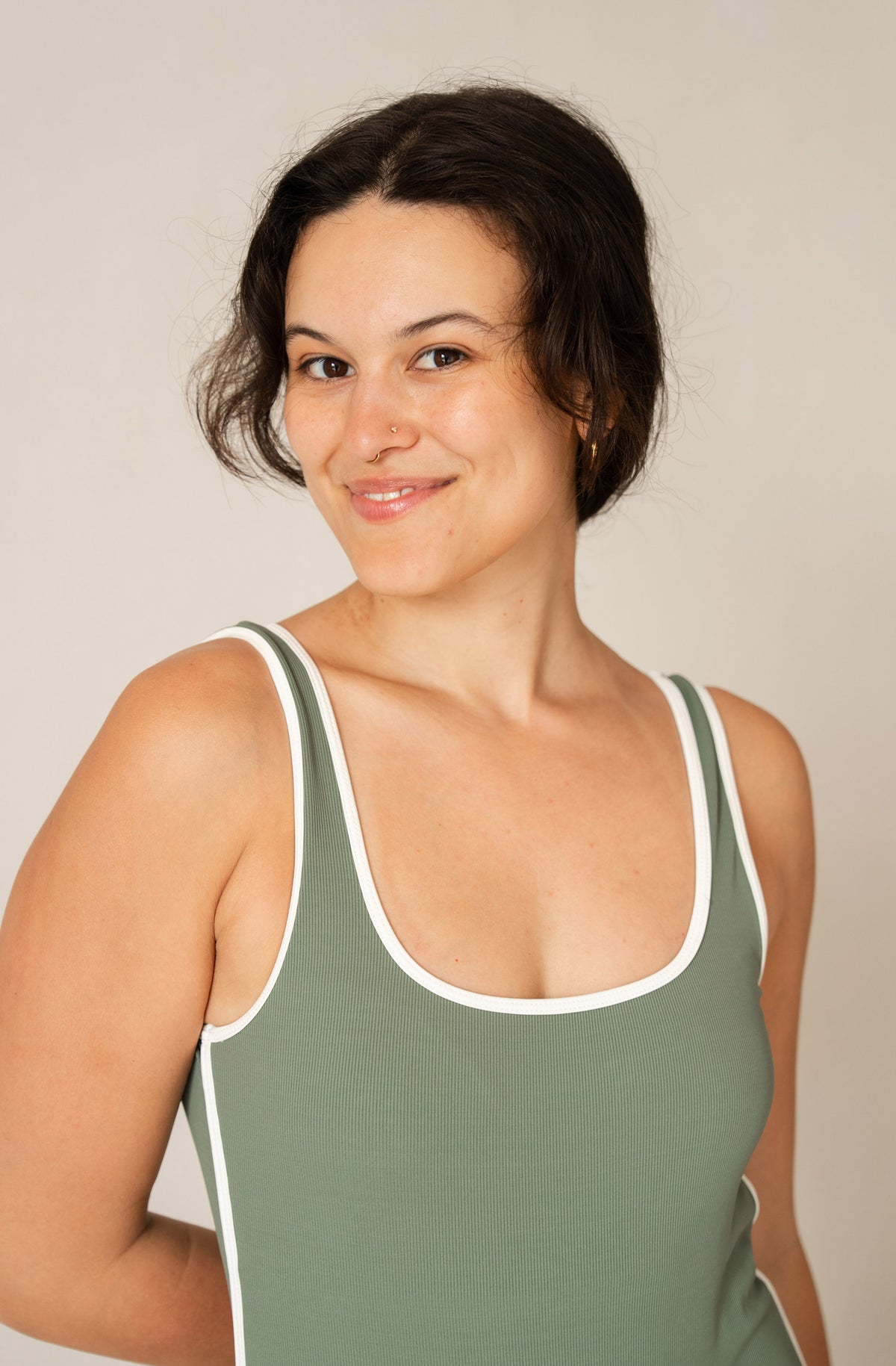 A woman with dark hair tied back wears a sage green tank top with white trim, photographed against a plain light beige studio background.