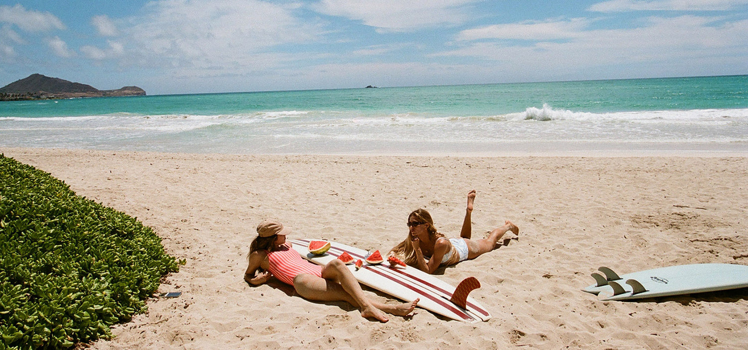 Two women relax on a red and white surfboard on a sandy beach, with turquoise ocean waves, a green hedge, and a second surfboard nearby.