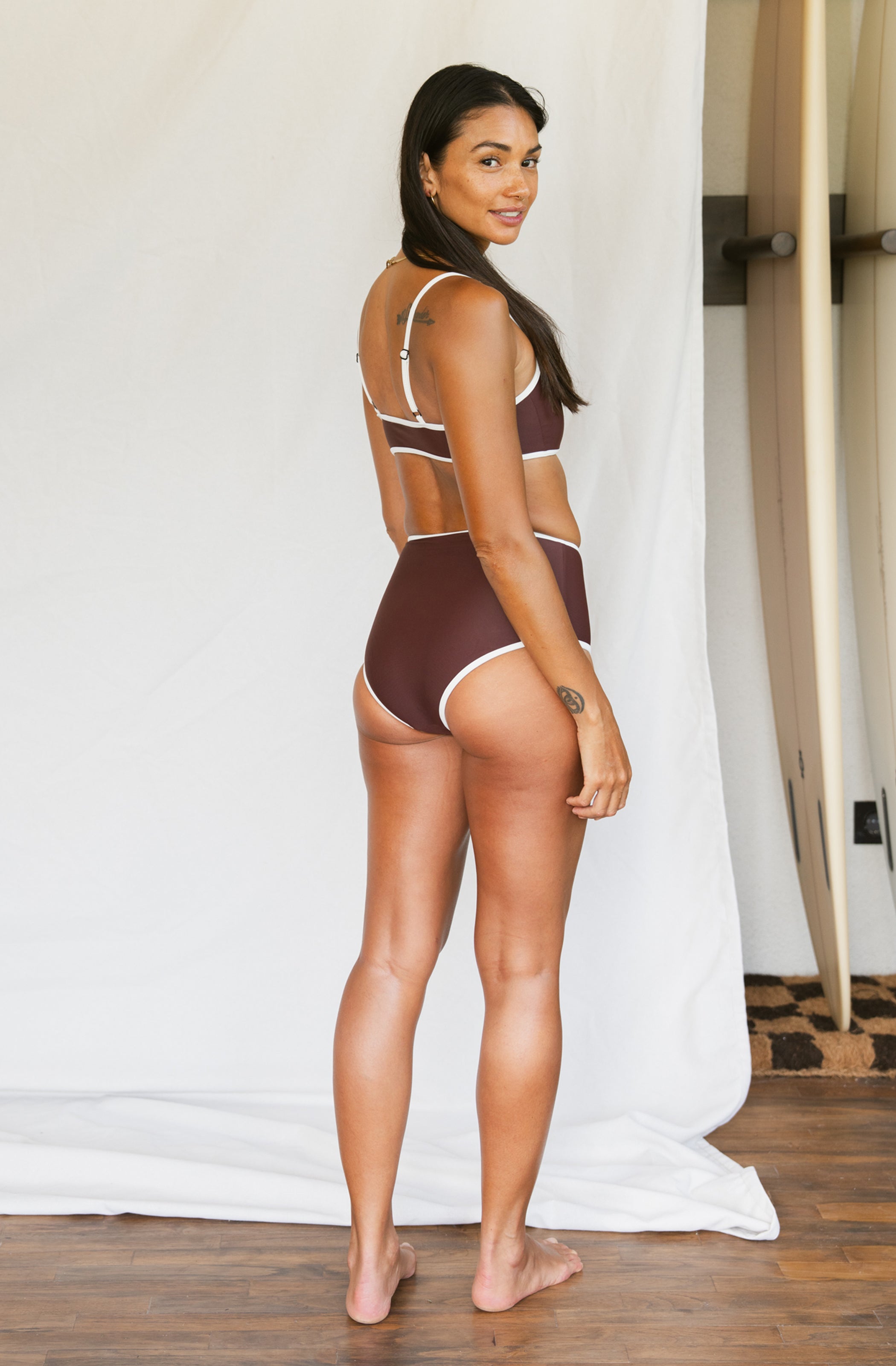 Woman wearing a brown bikini set standing against a white curtain.