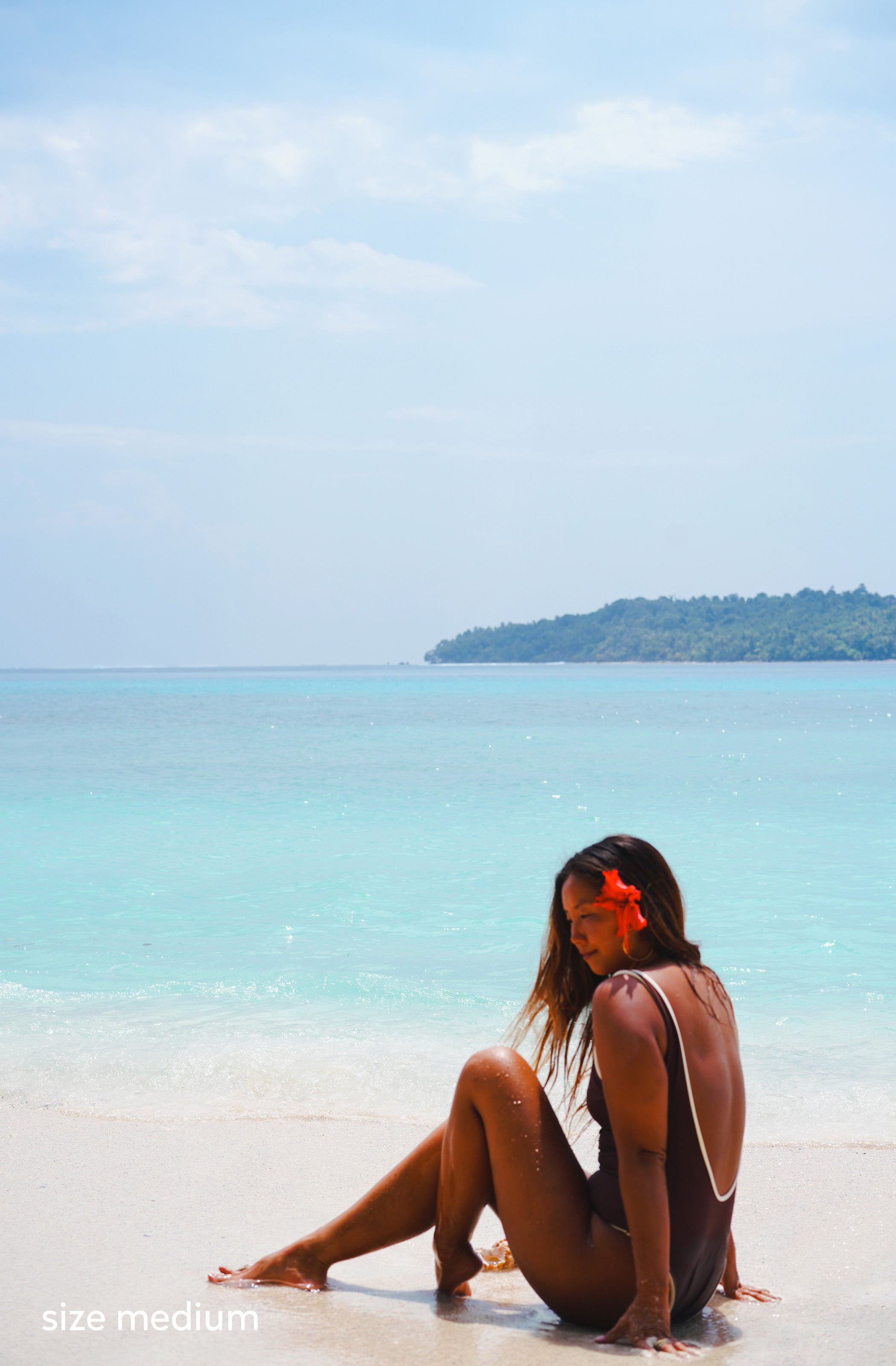 A woman standing on a beach wearing a one-piece swimsuit in brown with thin white trim. She has a red flower in her hair and is wearing gold hoop earrings.