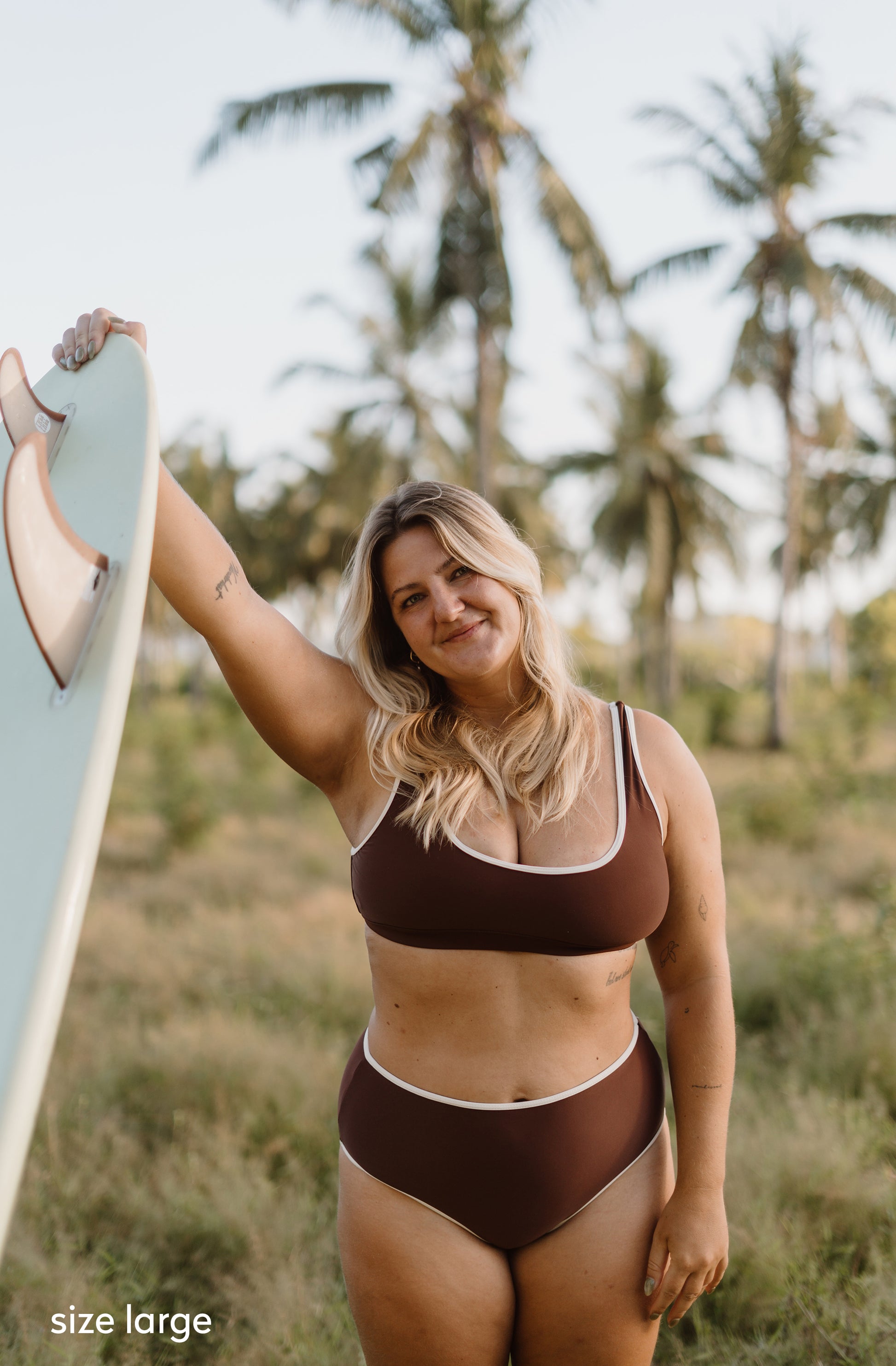 A woman wearing a brown two-piece bikini with white trim stands outdoors in a grassy area with palm trees, holding a light blue surfboard. The text 'size large' appears in the bottom-left corner.