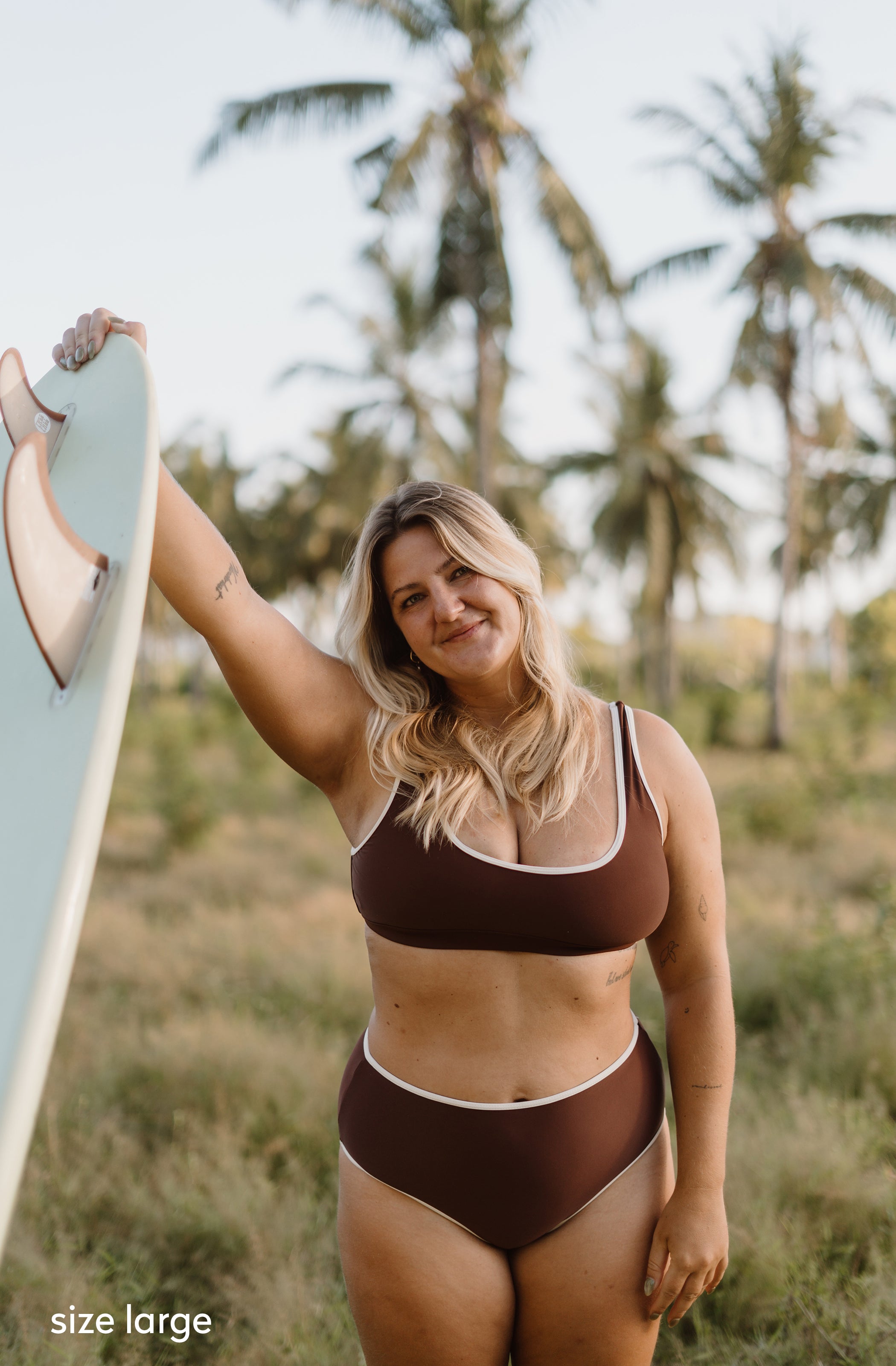A woman wearing a brown two-piece bikini with white trim stands outdoors in a grassy area with palm trees, holding a light blue surfboard. The text 'size large' appears in the bottom-left corner.