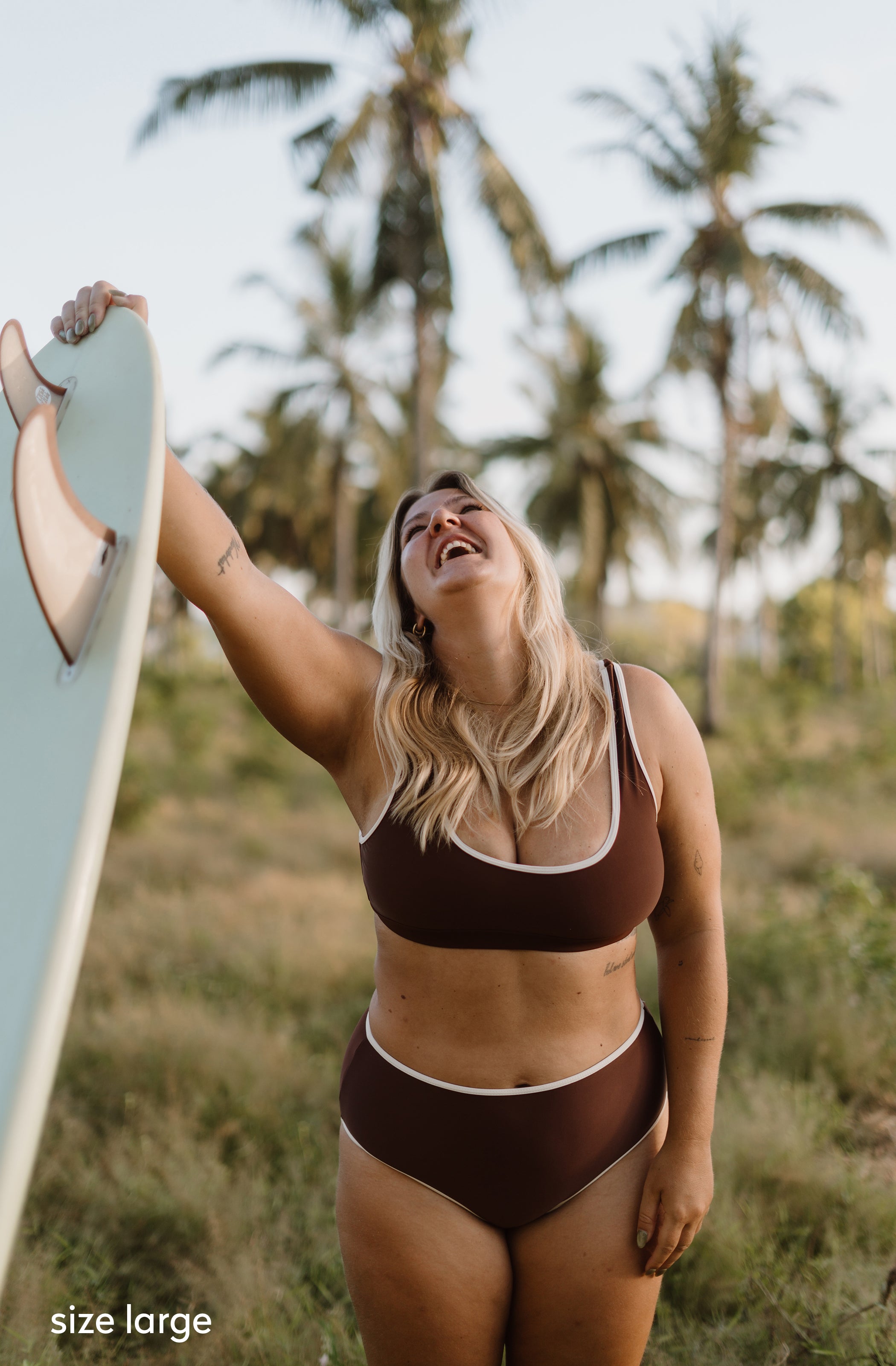 A woman outdoors wearing a brown two-piece bikini with white trim, holding a light blue surfboard, with palm trees in the background. A 'size large' text overlay is visible in the bottom-left corner.