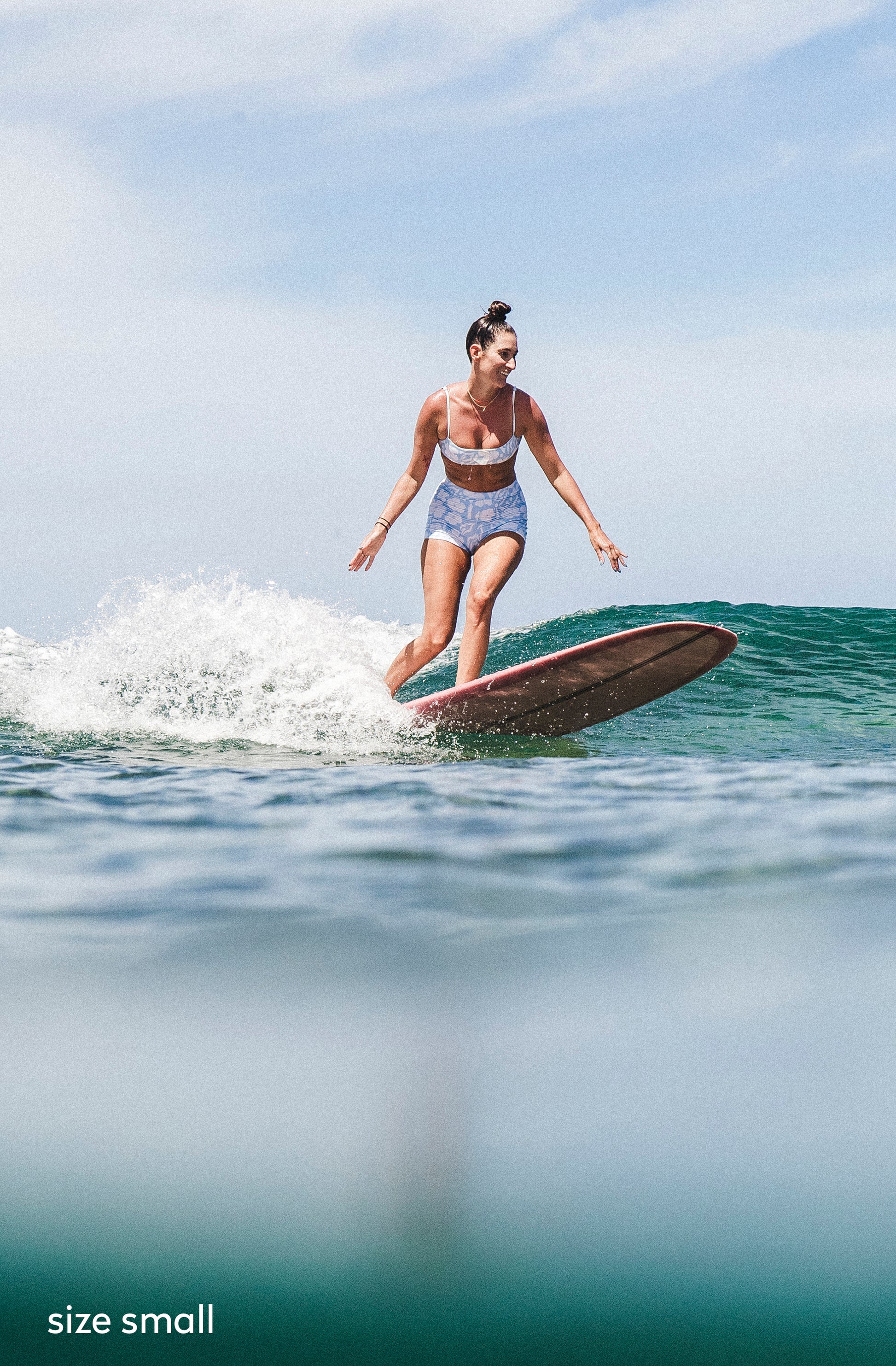 A woman stands on a surfboard in the ocean, wearing a blue bikini with a subtle blue pattern. The water is a deep teal-green, and the sky is light blue with light clouds.