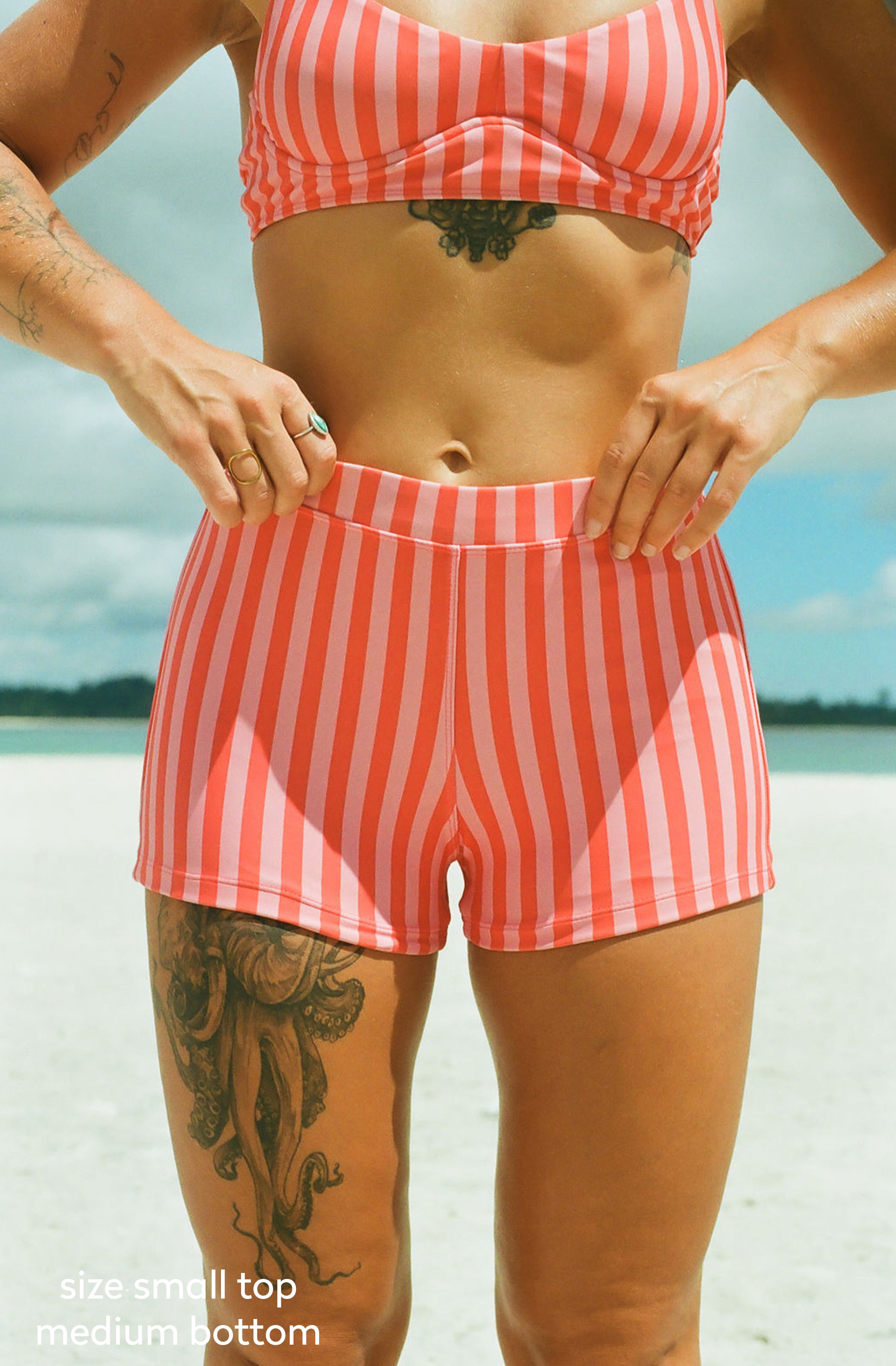 A woman wearing a coral and white striped bikini top and matching striped swim shorts on a beach. The photo is cropped from the chest down, with her hands on the waistband of the shorts.